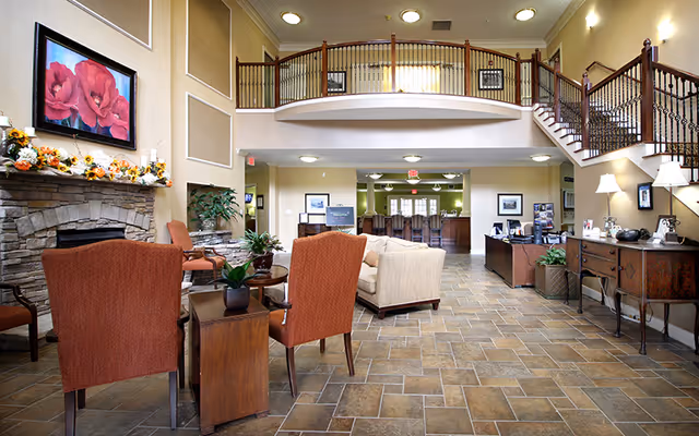 Spacious two-story senior living lobby with a seating area, stone fireplace, reception desk, and a mezzanine balcony.