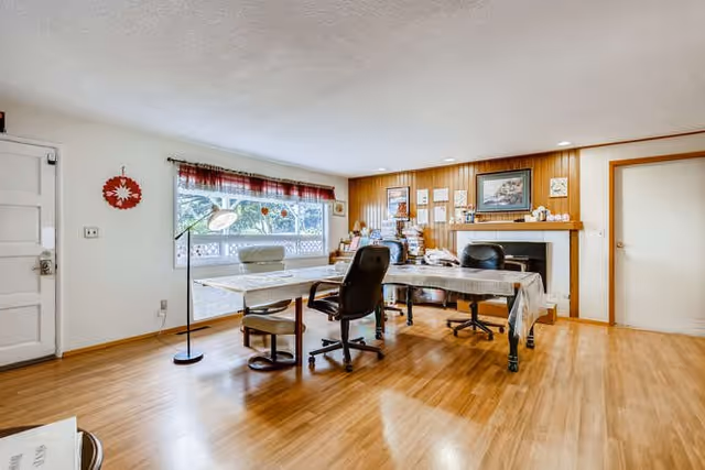 A spacious room with wooden flooring featuring two tables covered with tablecloths and surrounded by office chairs. There is a large window with a red valance letting in natural light, a white door on the left, and a fireplace with a wooden mantel on the right wall. Various framed pictures and decorations are displayed above the fireplace.