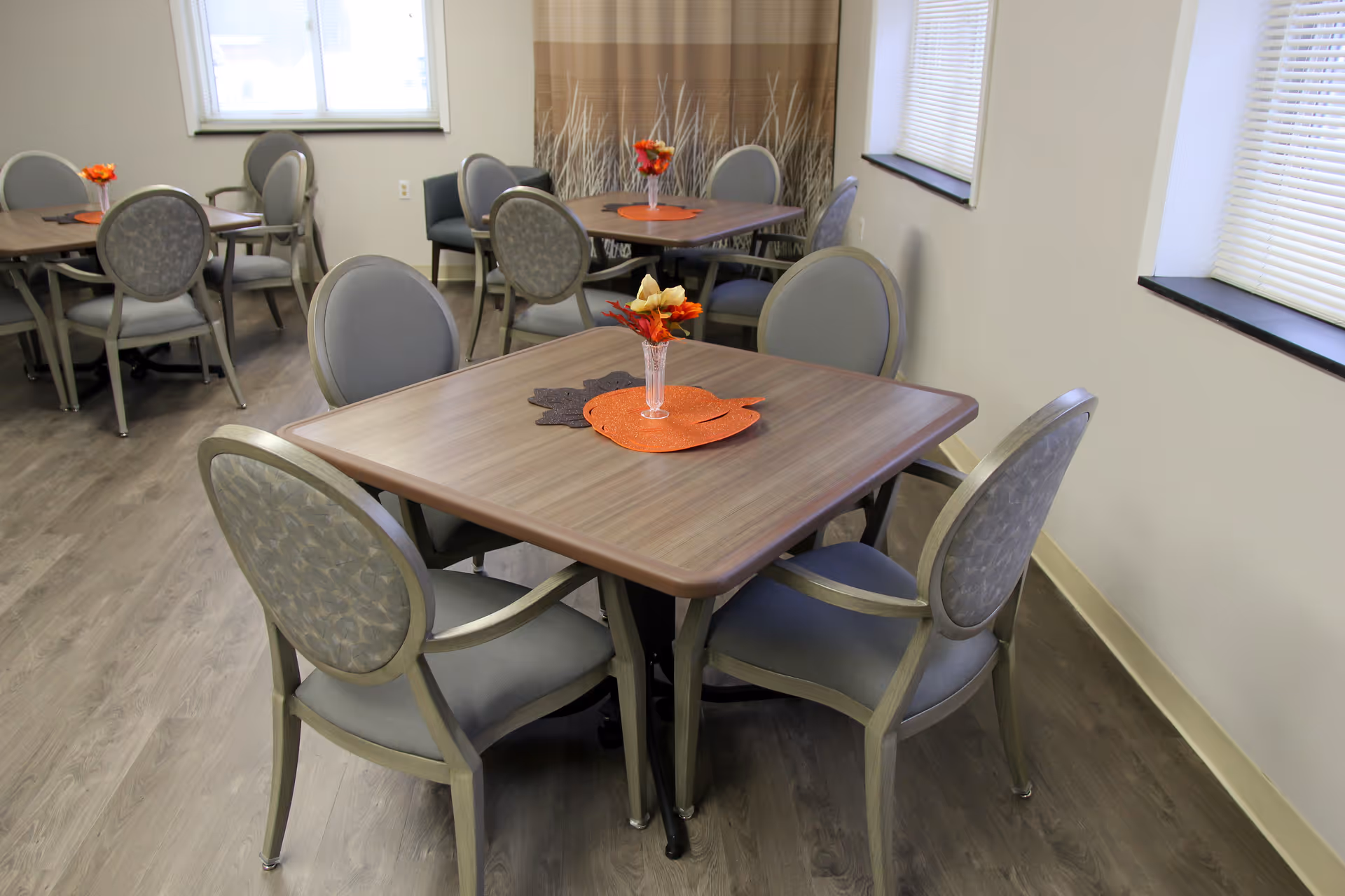 A dining area with multiple square wooden tables, each surrounded by four cushioned chairs with patterned backs. Each table has a small vase with artificial flowers and a decorative placemat. The room has light-colored walls, windows with blinds, and wood-look flooring.