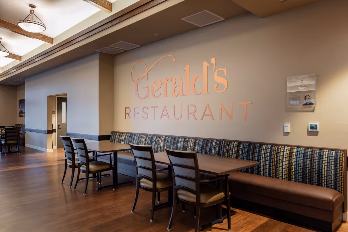 Interior view of Gerald's Restaurant featuring a long cushioned bench with patterned upholstery along the wall, wooden tables and chairs, warm lighting, and wooden ceiling beams.