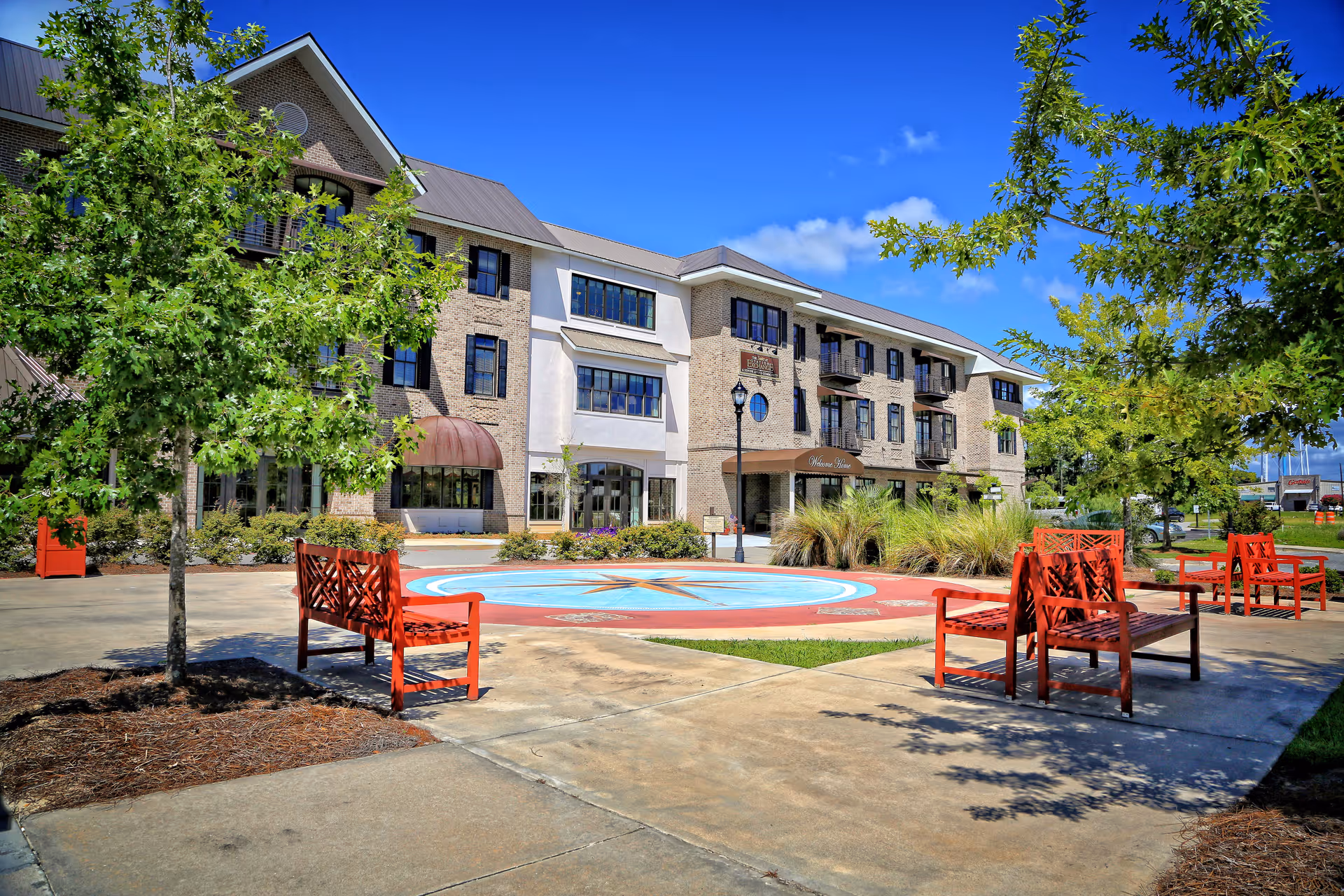 Outdoor courtyard area of Station Exchange Senior Living featuring red benches, trees, a circular compass design on the ground, and a multi-story brick and stucco building under a clear blue sky.