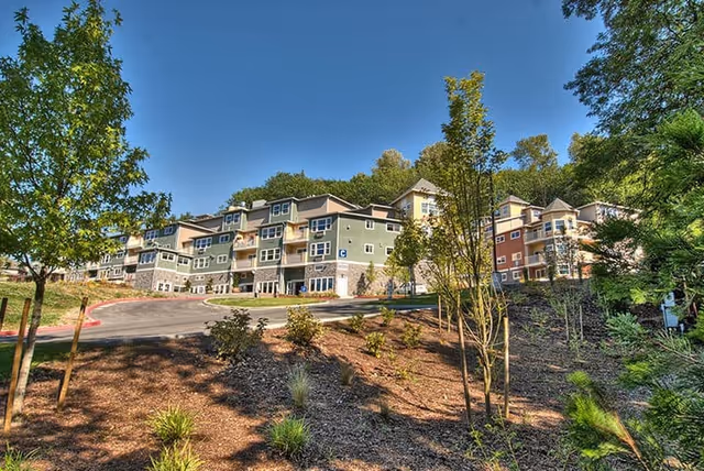 Exterior view of a multi-story senior living facility building surrounded by landscaped grounds with trees and shrubs under a clear blue sky.