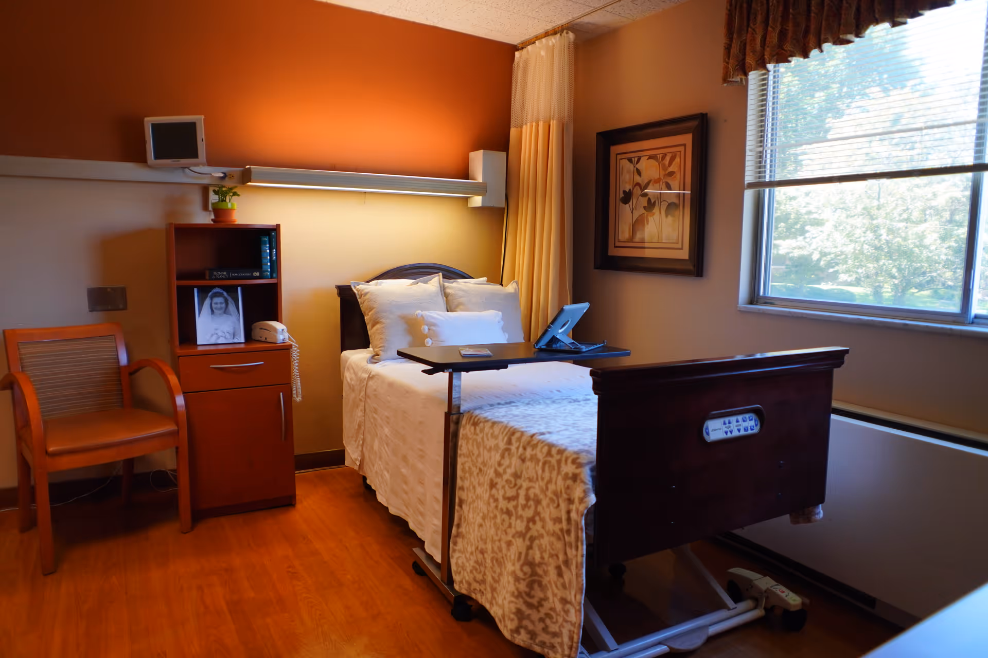 A furnished assisted-living bedroom with a single adjustable bed, overbed table, nightstand with phone and framed photo, chair, and a window.