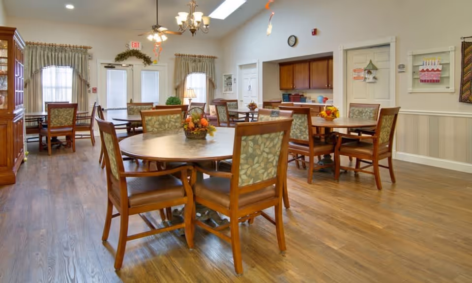 A bright and spacious dining room in a senior living facility with several round wooden tables surrounded by cushioned chairs. The room features wood flooring, large windows with curtains, a chandelier and ceiling fan, and a serving area with cabinets. Decorative items and a clock are mounted on the walls.