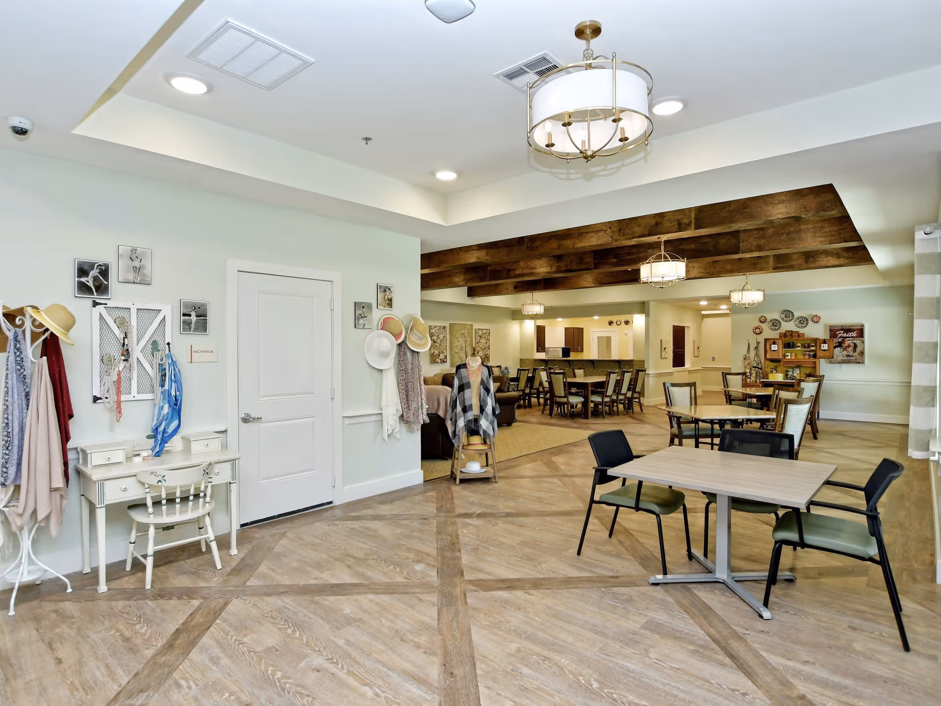 A spacious and well-lit common area in a senior living facility named The Philomena, featuring wooden flooring, multiple tables and chairs, decorative hats and scarves on a coat rack and wall hooks, a small white desk with a chair, and wooden beams on the ceiling. The room has soft lighting from ceiling fixtures and a cozy, inviting atmosphere.