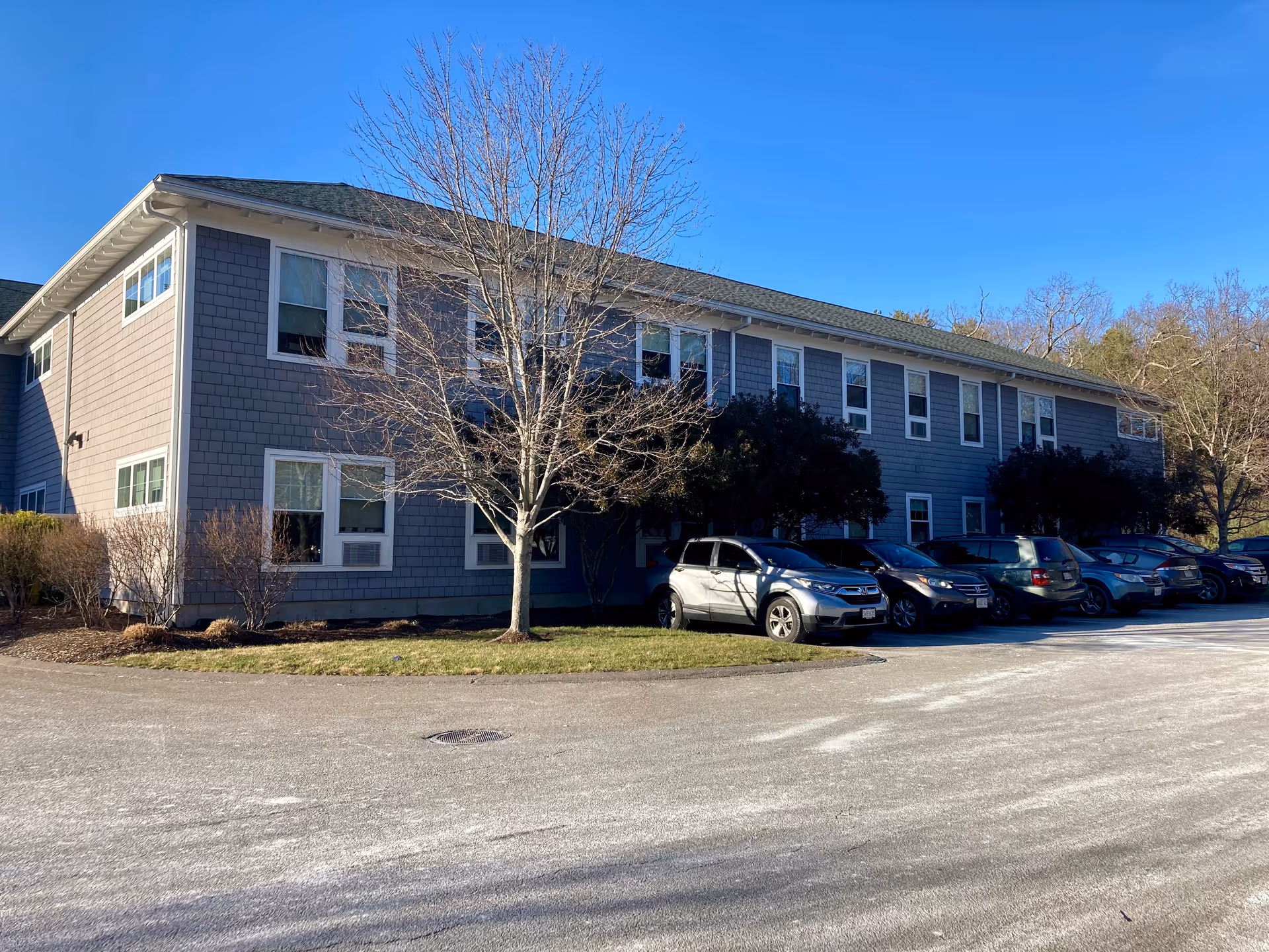 Exterior view of a two-story gray building with multiple windows, a leafless tree, bushes, and a parking lot with several parked cars under a clear blue sky.