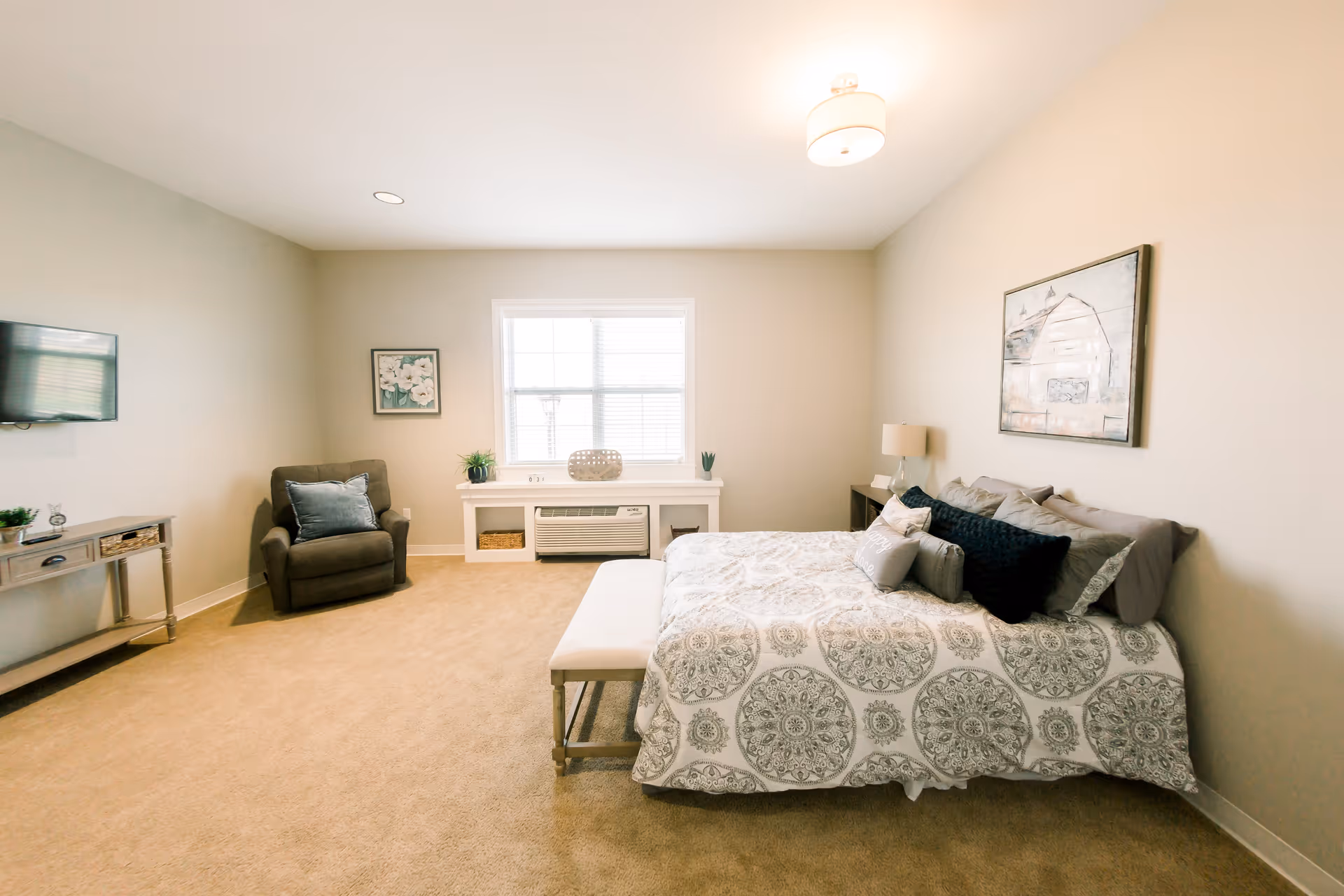 A spacious bedroom with a large bed covered in patterned bedding and multiple pillows. There is a bench at the foot of the bed, a comfortable armchair with a cushion, a wall-mounted TV, a small console table, and a window with blinds letting in natural light. The walls are painted a light beige color and there is a piece of artwork hanging above the bed.