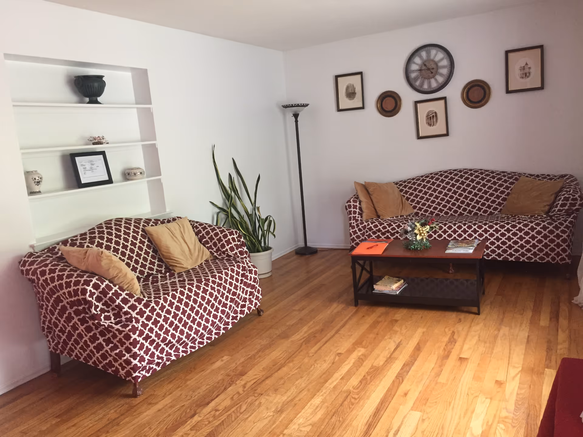 A living room with two patterned sofas covered in maroon and white fabric, each with beige throw pillows. A wooden coffee table with magazines and a small decorative plant sits between the sofas. The room has hardwood floors, a tall floor lamp, a potted plant, and framed artwork and a clock on the wall.