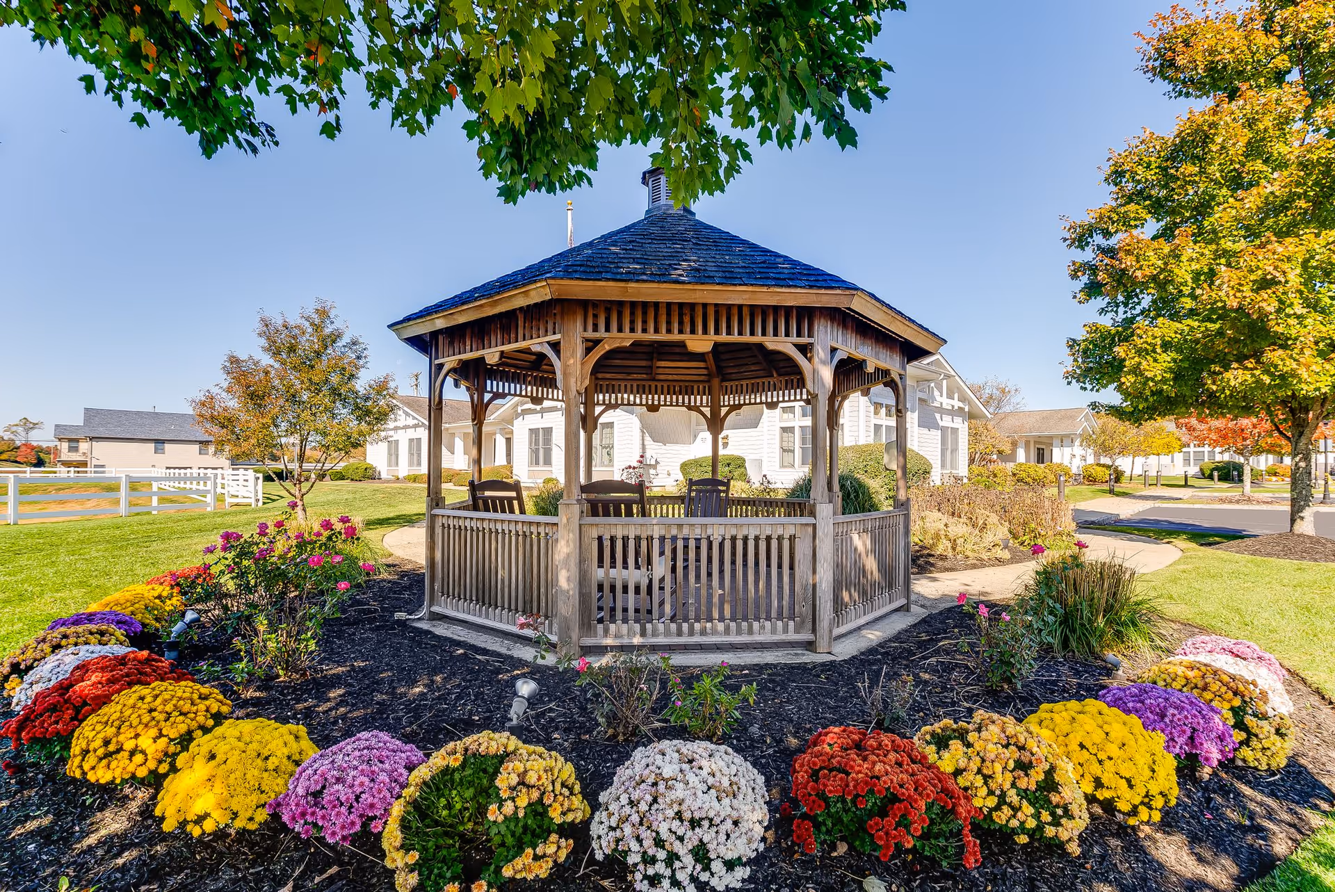 A wooden gazebo surrounded by colorful flowers and greenery in a well-maintained garden area with a clear blue sky and buildings in the background.