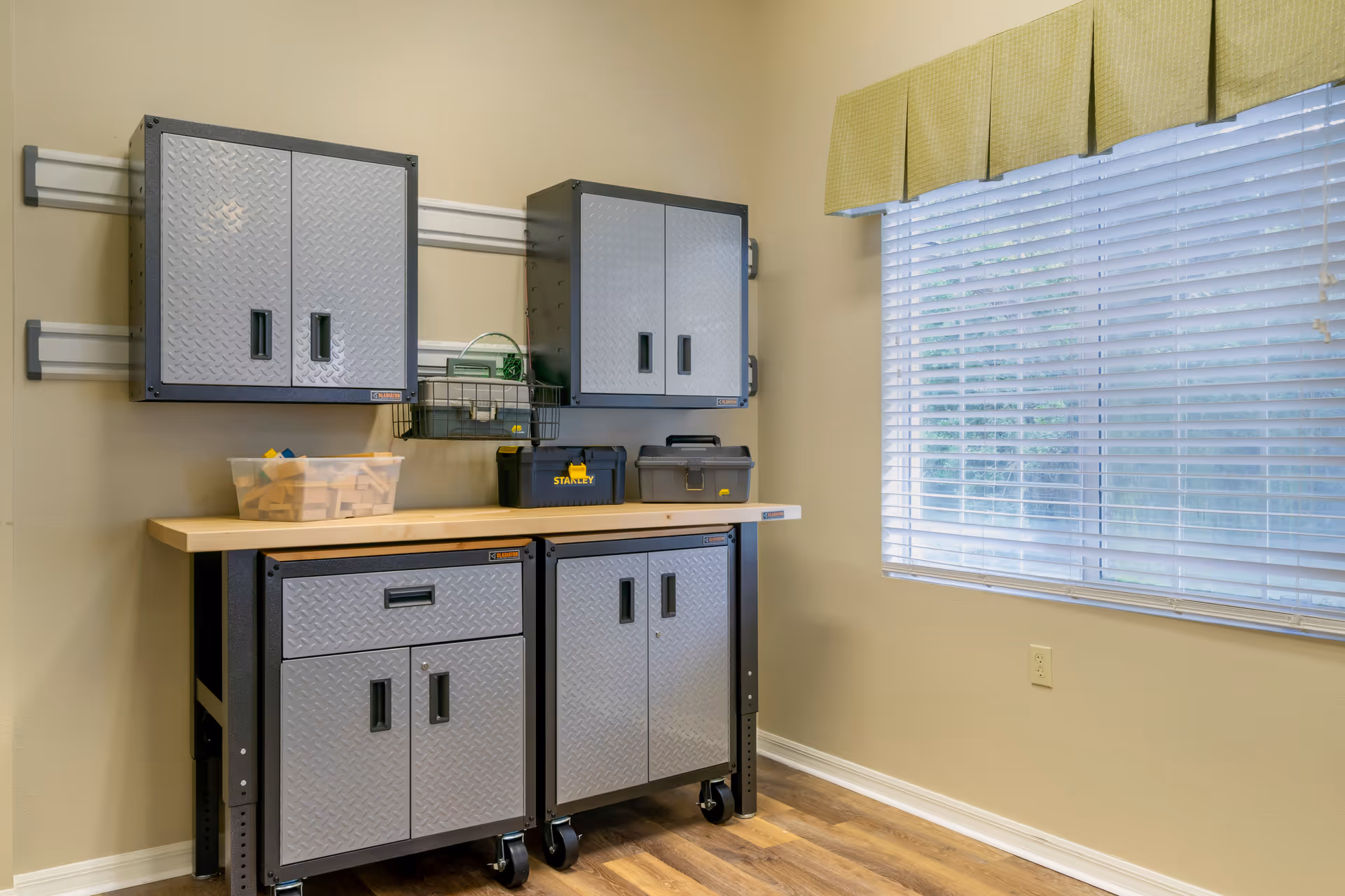 A small utility room featuring gray metal wall cabinets and a wheeled workbench with toolboxes beneath a window with blinds and a green valance.