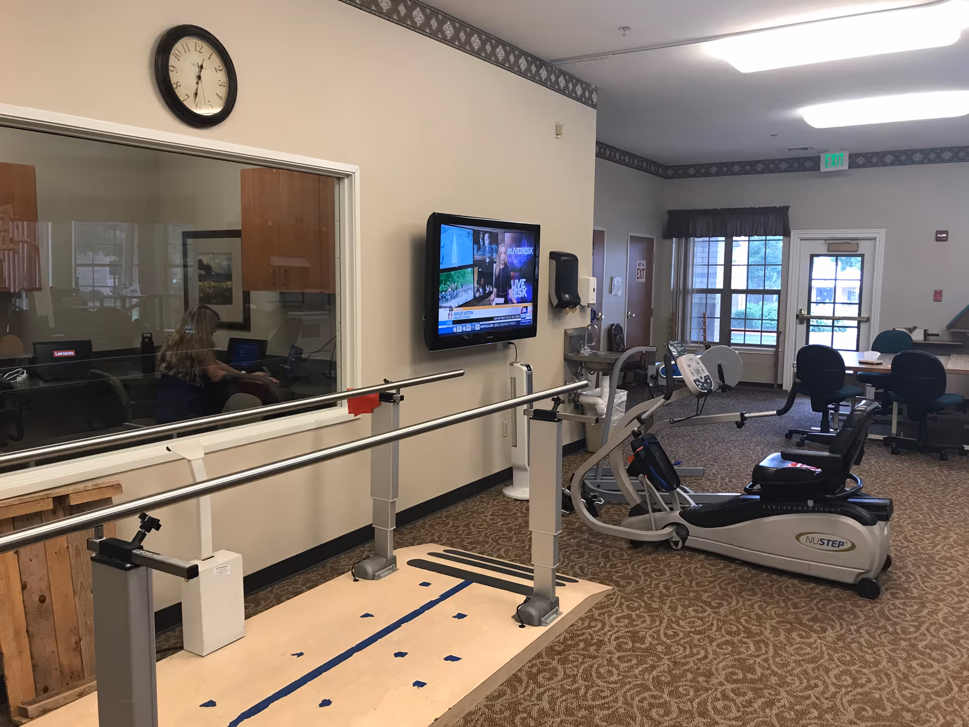 Interior view of a rehabilitation or therapy room in a senior living facility. The room features parallel bars for walking exercises, a NuStep recumbent cross trainer, a wall-mounted TV, a clock, and a window into an adjacent office where a person is working on a laptop. There are tables and chairs in the background near a door and windows with curtains.