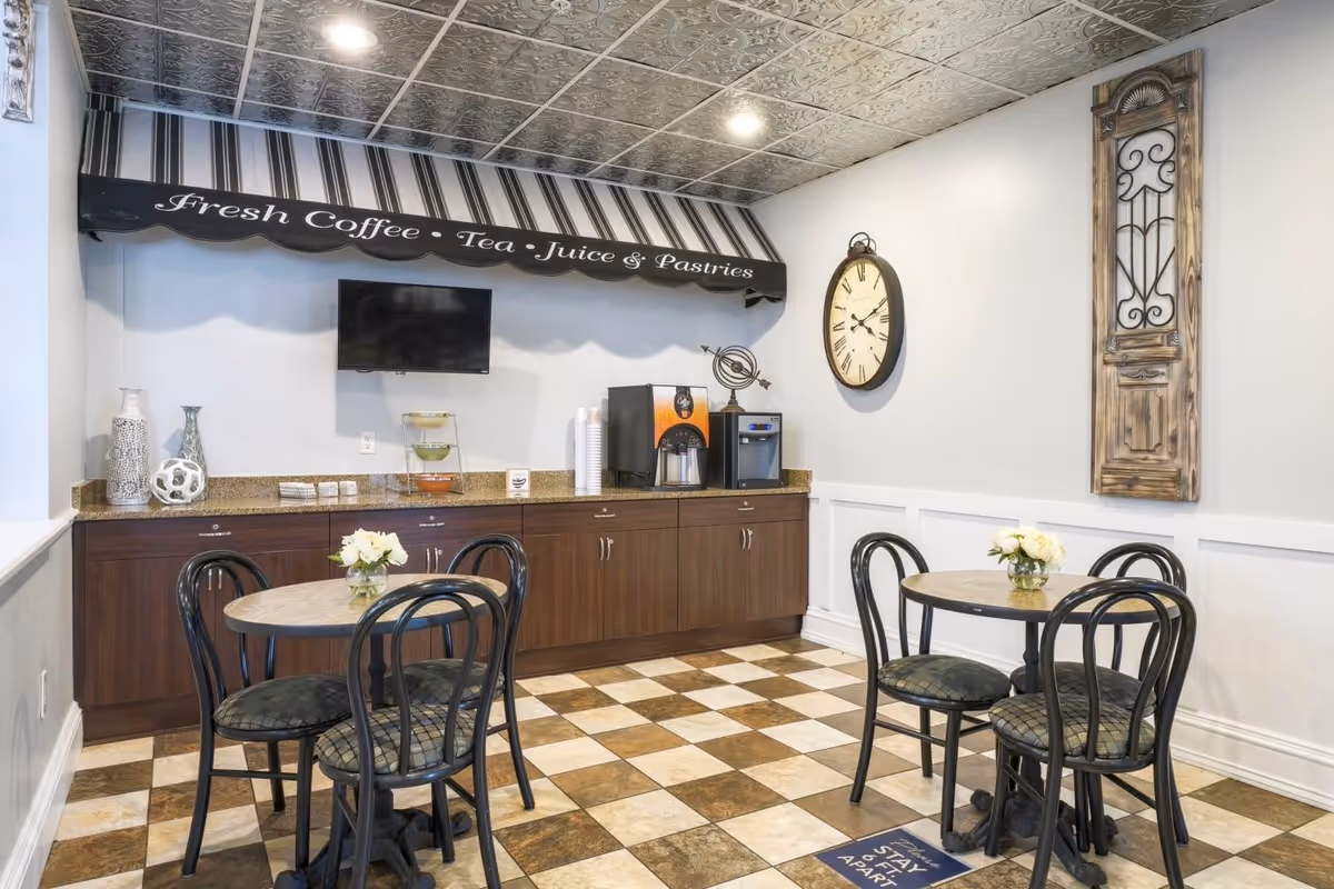A cozy dining area with two round tables, each surrounded by four black chairs with patterned cushions. The floor has a checkered tile pattern in brown and beige. Against the wall is a countertop with a coffee machine, cups, and decorative vases. Above the counter is a striped awning with the text 'Fresh Coffee • Tea • Juice & Pastries'. A large wall clock and a decorative wooden panel hang on the adjacent wall.