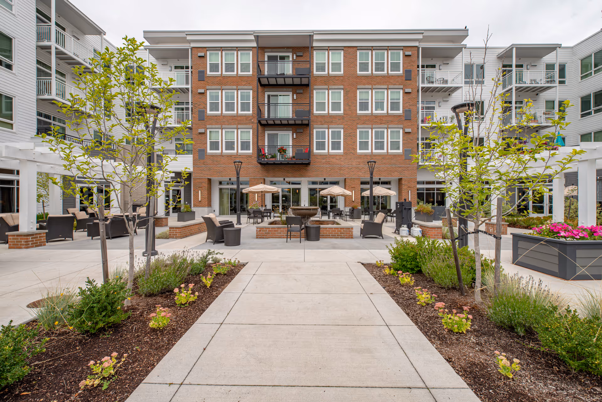 Courtyard patio with seating, umbrellas, and landscaping in front of a multi-story brick and siding senior living building.