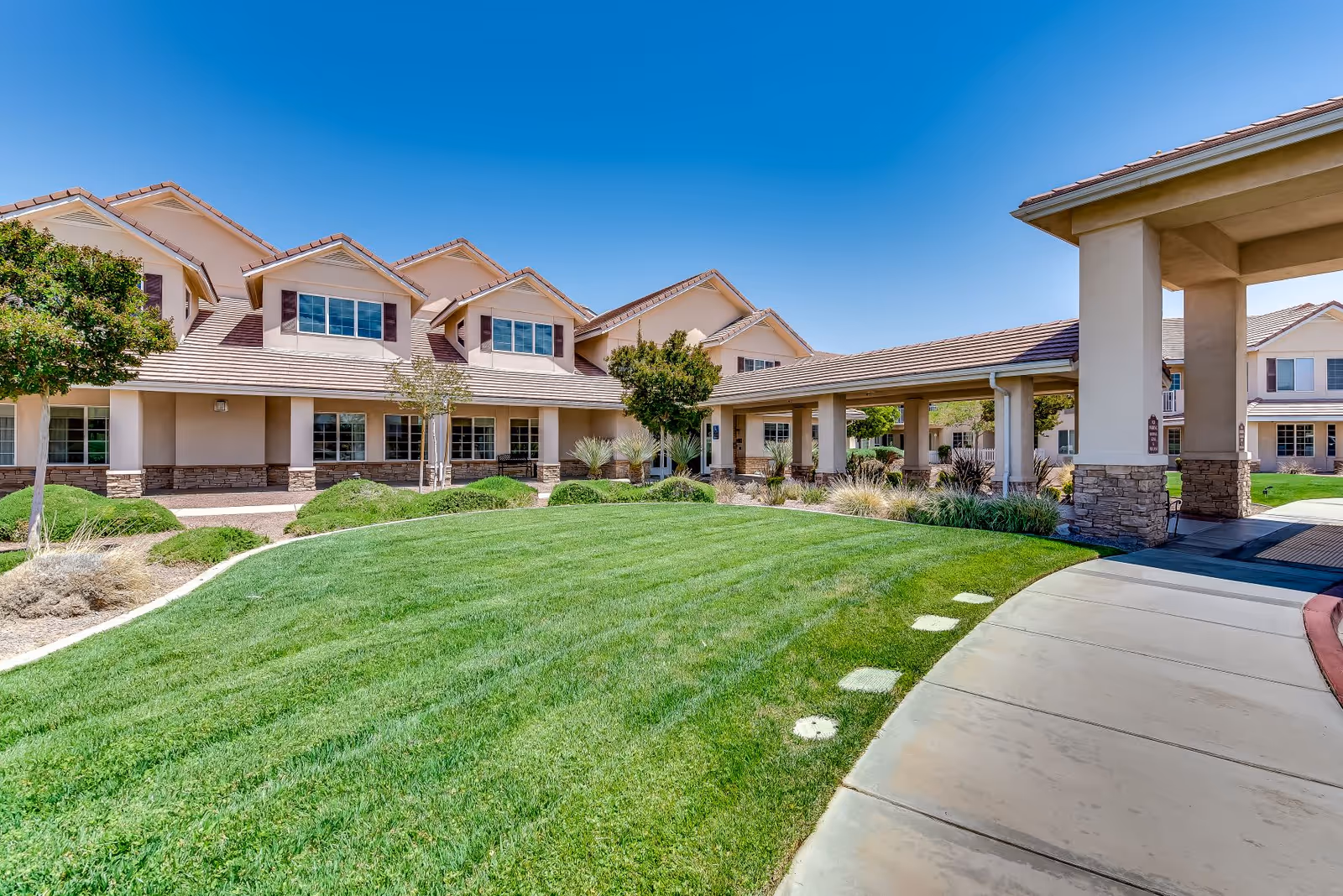 Exterior view of a senior living facility building with beige walls, multiple windows, and a tiled roof under a clear blue sky. The foreground features a well-maintained green lawn, small trees, shrubs, and a curved concrete walkway leading to a covered entrance supported by columns.