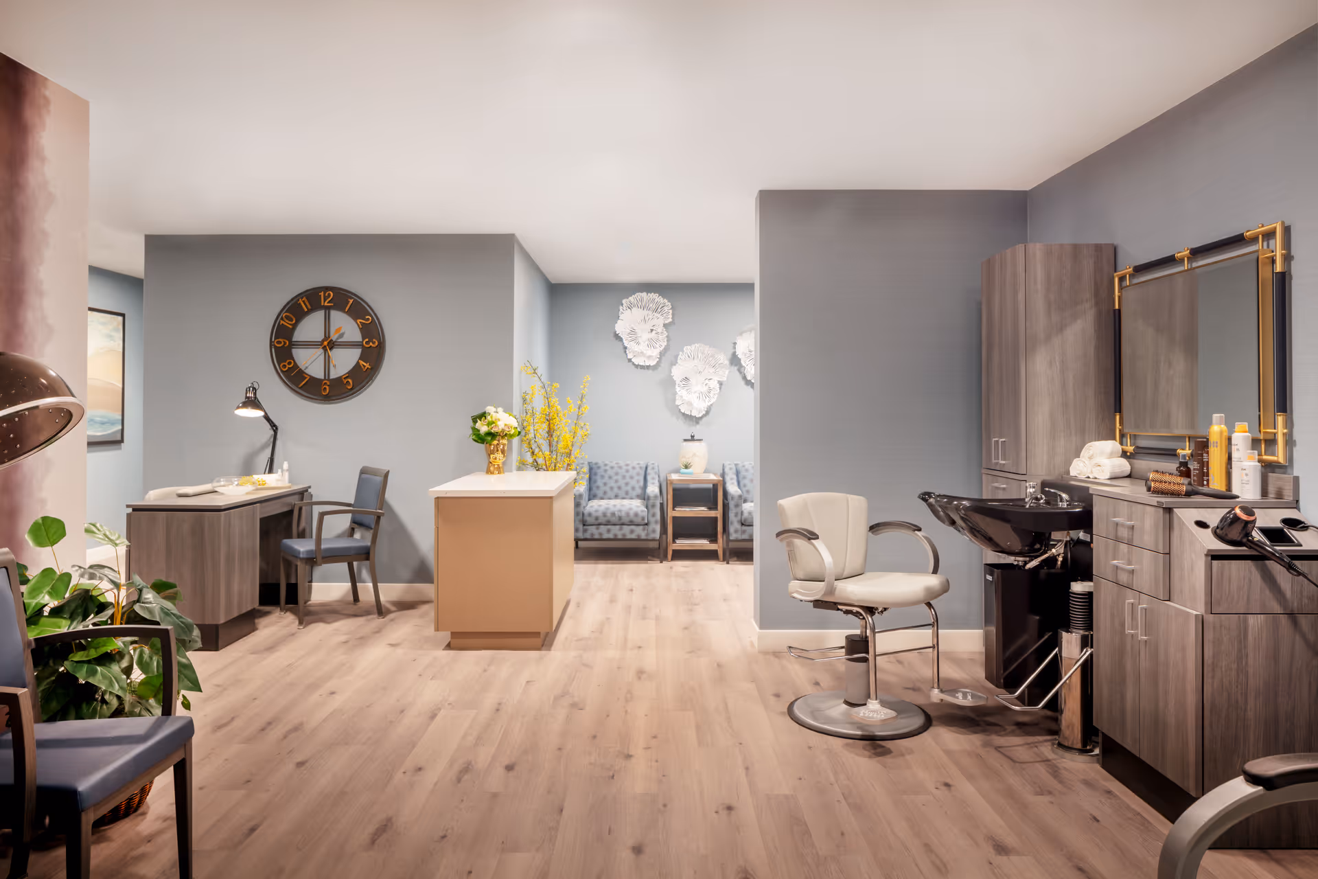 A modern and clean salon area within a senior living facility featuring a styling chair and wash basin on the right, a desk with a chair and lamp on the left, a central island with flowers, and a seating area with two armchairs and decorative wall art in the background. The room has light wood flooring and soft gray walls.