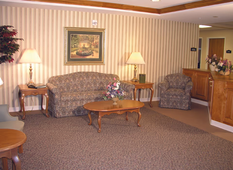 Decorative sitting area with a patterned sofa and armchair, wooden coffee and side tables, lamps, floral arrangements, and framed artwork on striped wallpaper.