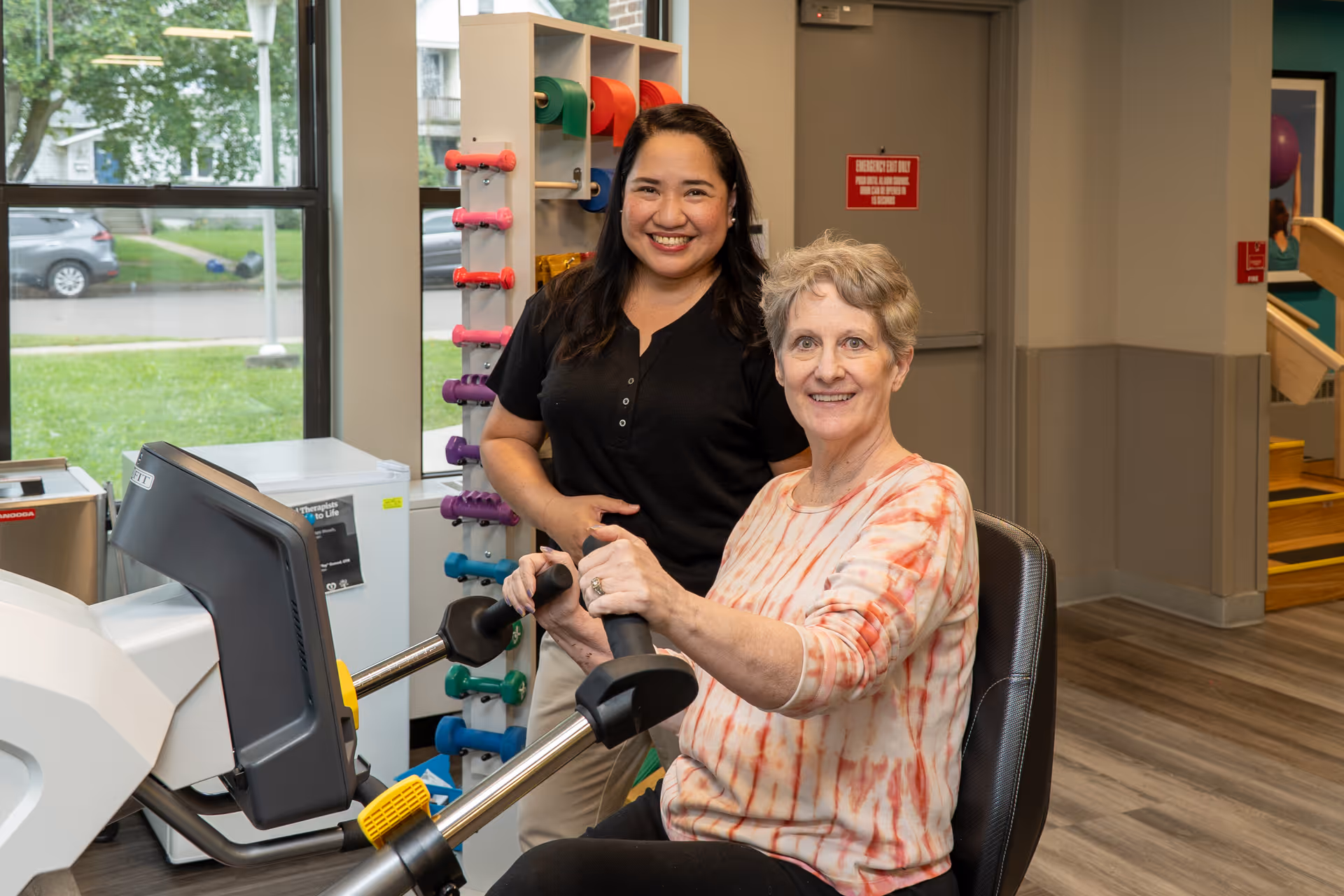 A senior woman using a seated exercise machine in a rehabilitation center, assisted by a smiling female staff member. Colorful dumbbells are visible on a rack behind them, and a large window shows a green outdoor area with a parked car.