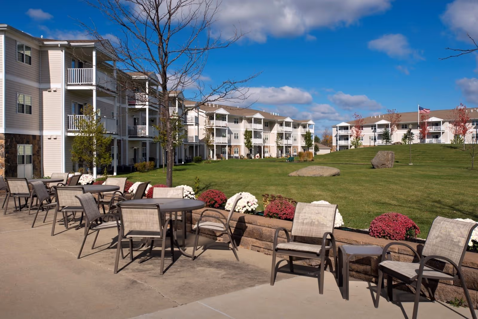 Outdoor patio area with multiple tables and chairs arranged on a concrete surface next to a landscaped grassy area with flowers and trees. In the background, there are several three-story residential buildings under a blue sky with some clouds.