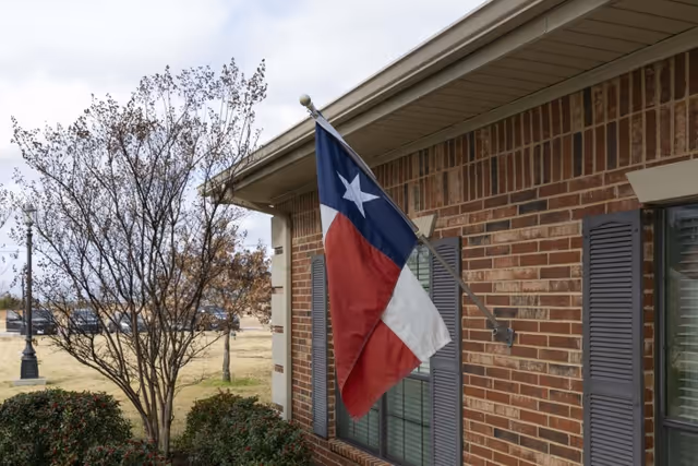 A Texas flag flies from a pole mounted on the brick exterior of a building with windows, shutters, shrubs, and a leafless tree.