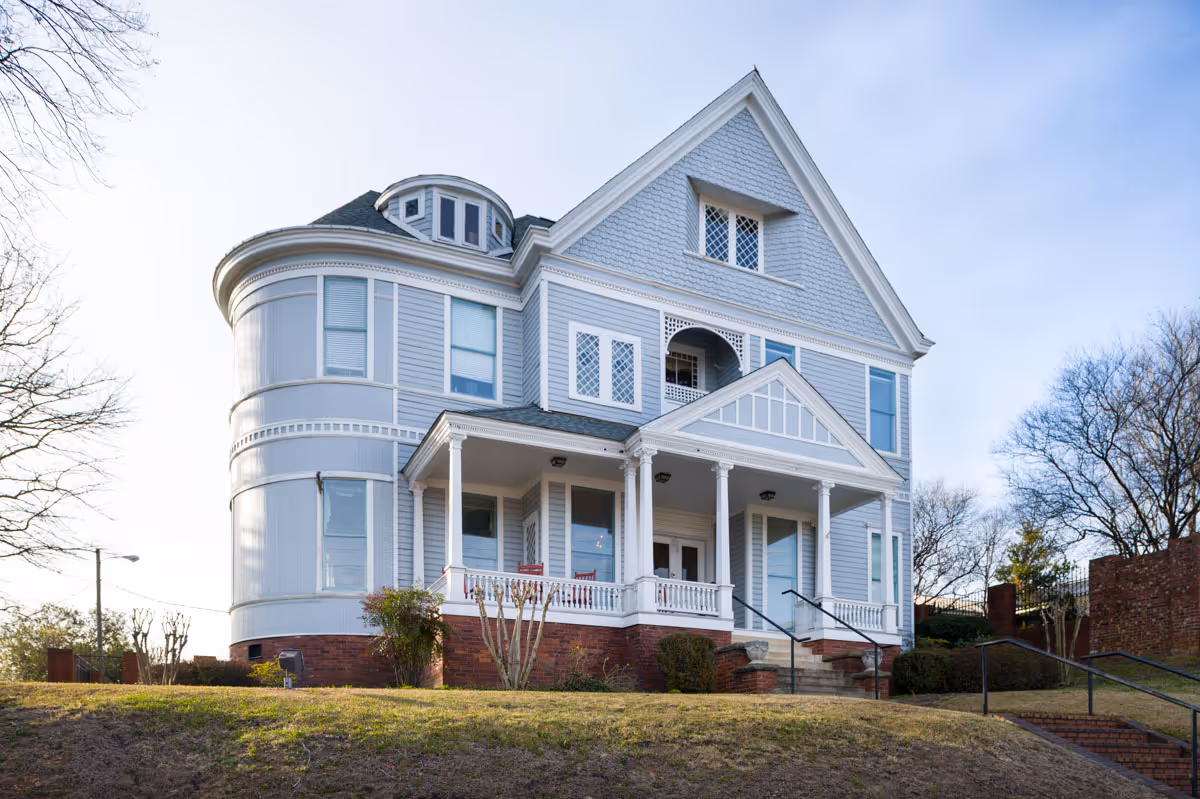 Exterior view of a large, light blue Victorian-style house with white trim, a front porch with columns, and multiple windows. The house is situated on a grassy hill with some bushes and trees around it under a clear sky.