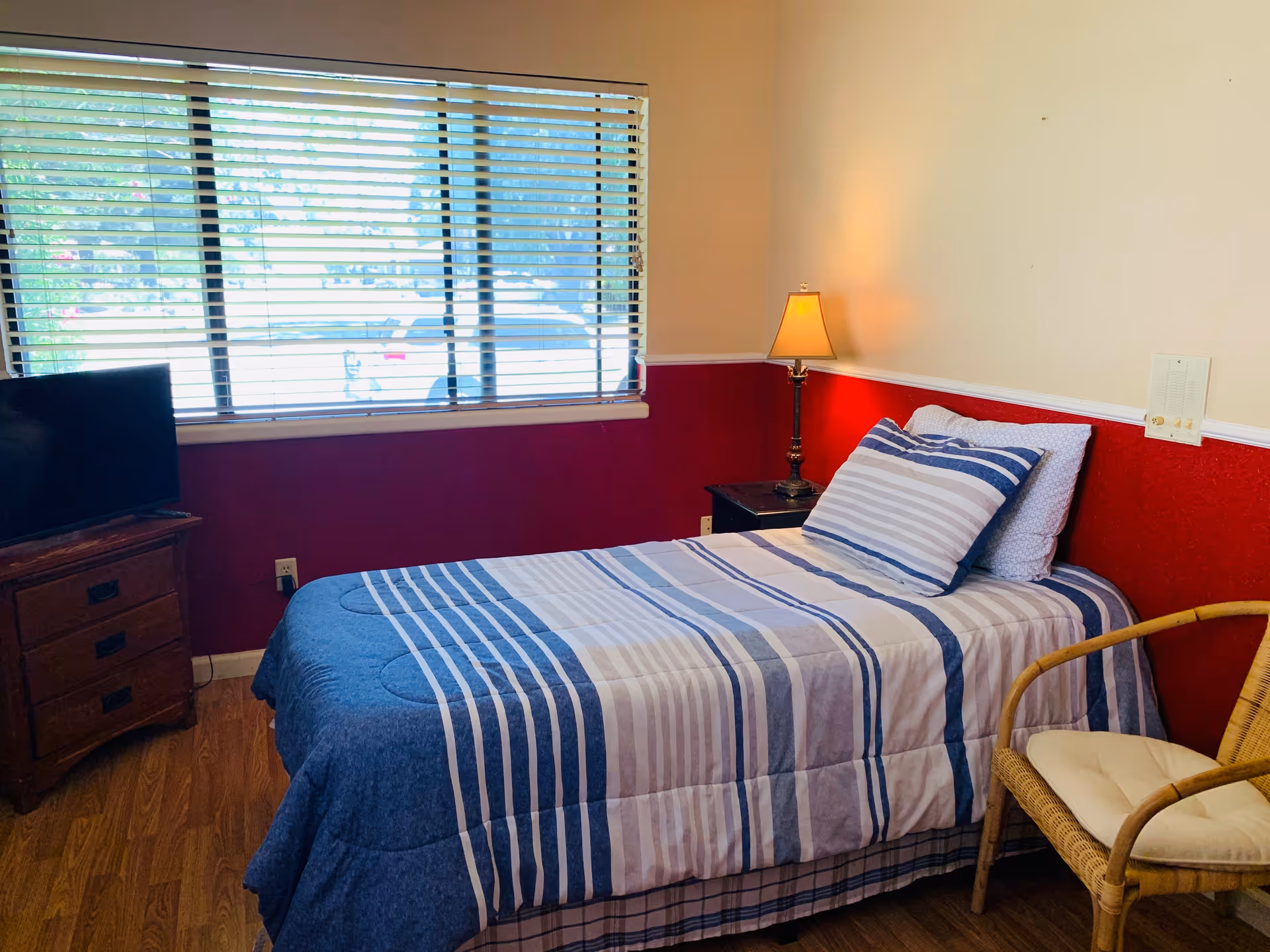 A small bedroom with a single bed covered in blue and white striped bedding. There is a wooden nightstand with a lamp on it next to the bed, a wicker chair with a cushion, and a wooden dresser with a flat-screen TV on top. A large window with horizontal blinds lets in natural light.