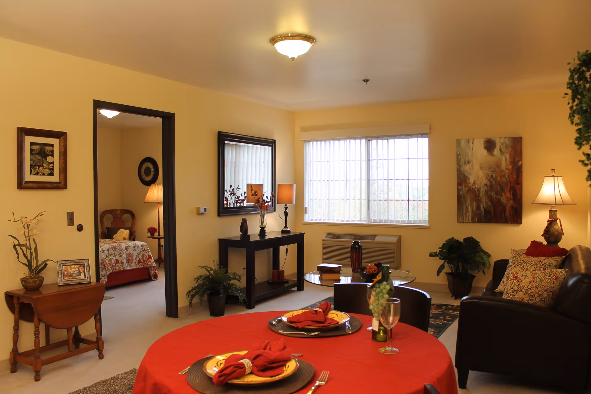 A cozy living room and dining area in a senior living facility with a round table covered with a red tablecloth set for two. The room has a black leather couch with patterned pillows, a small black coffee table, a console table with decorative items, and a large window with vertical blinds. A bedroom with a floral bedspread and a lamp is visible through an open doorway.