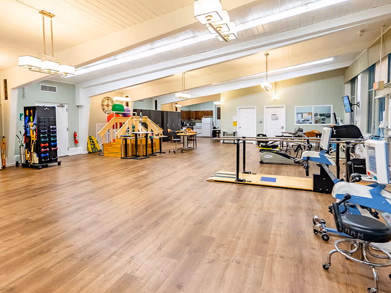 A spacious rehabilitation room with wooden flooring, equipped with parallel bars for walking exercises, exercise machines, a rack of colorful dumbbells, and a small set of stairs with handrails. The room has soft lighting from ceiling fixtures and large windows on the right side.