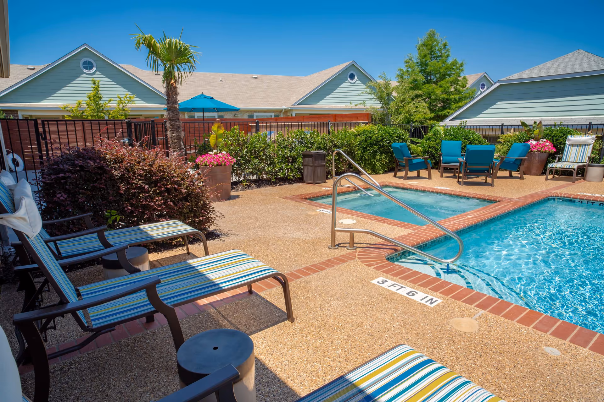 Outdoor pool area with clear blue water, surrounded by a concrete deck with striped lounge chairs and blue cushioned chairs. There are potted plants, a palm tree, and a fence enclosing the area. Residential-style buildings with light green siding and triangular roofs are visible in the background under a clear blue sky.