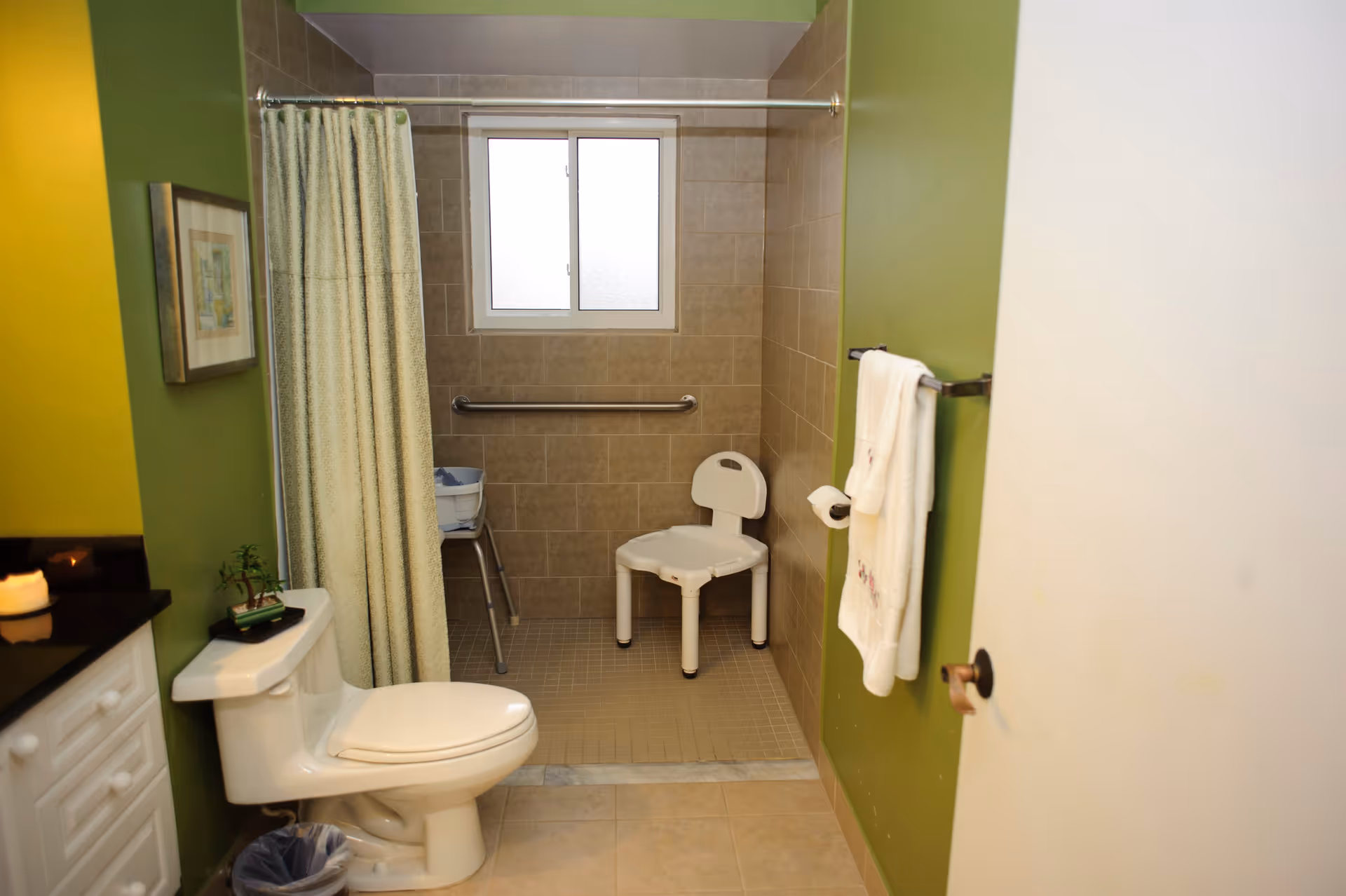 Bathroom with green walls featuring a toilet, a shower area with a white shower chair, a grab bar, a window, a towel hanging on a rack, and a countertop with a candle and small plant.