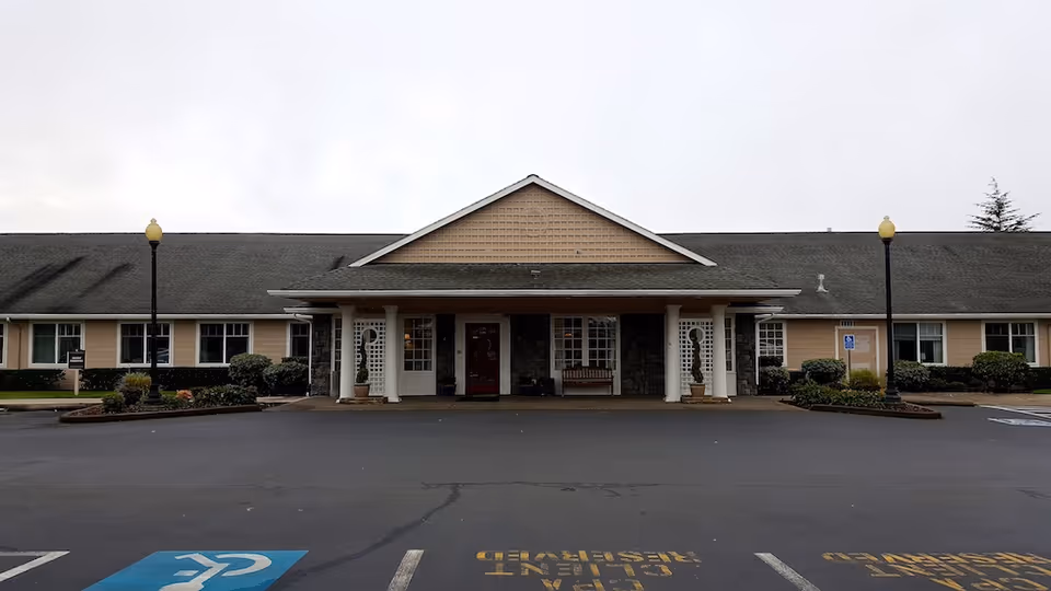 Front entrance of a single-story memory care building with a covered portico, columns, landscaping, and a parking area with marked handicap spaces.