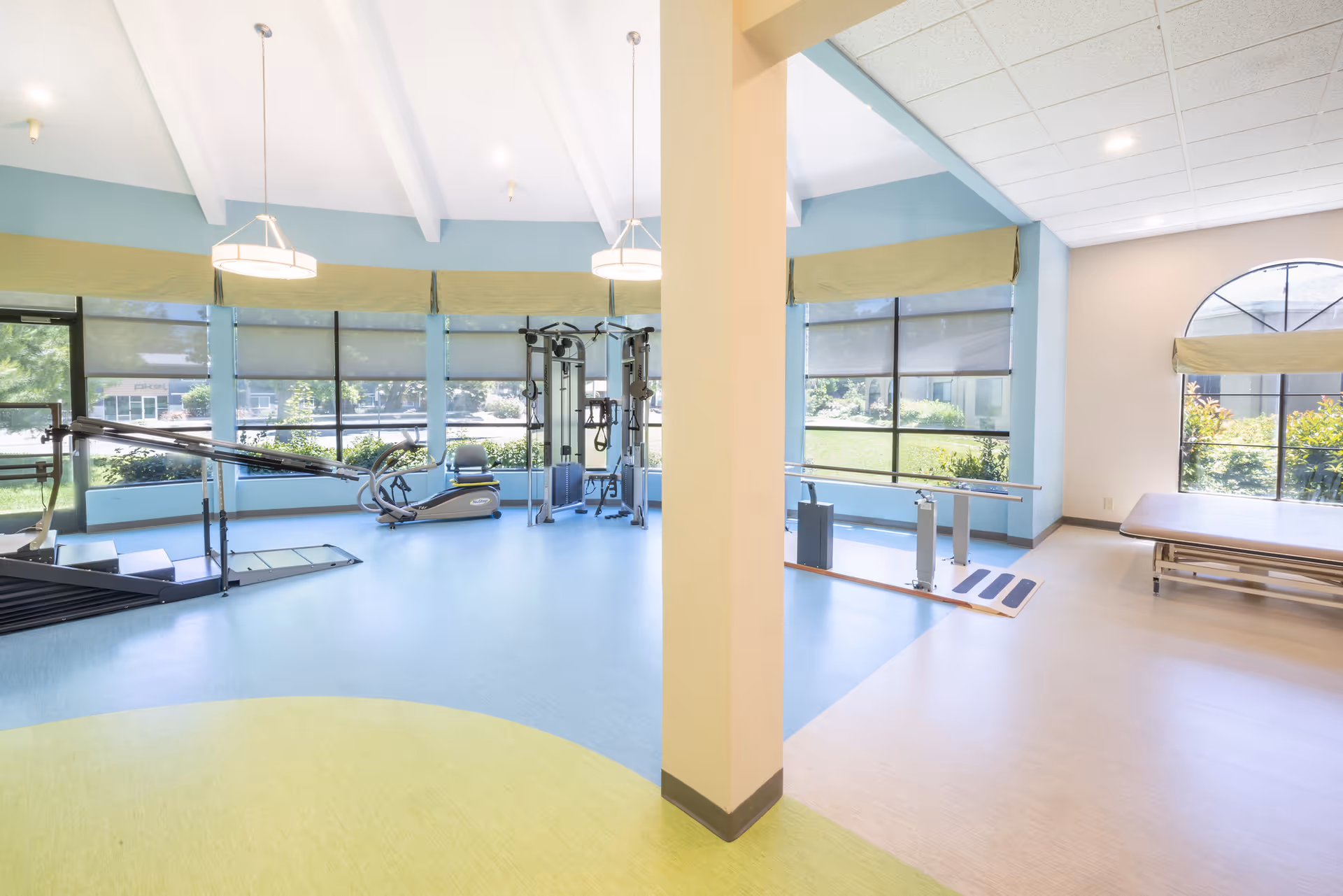 Bright rehabilitation gym with exercise machines, parallel bars, and a treatment table beside large windows.