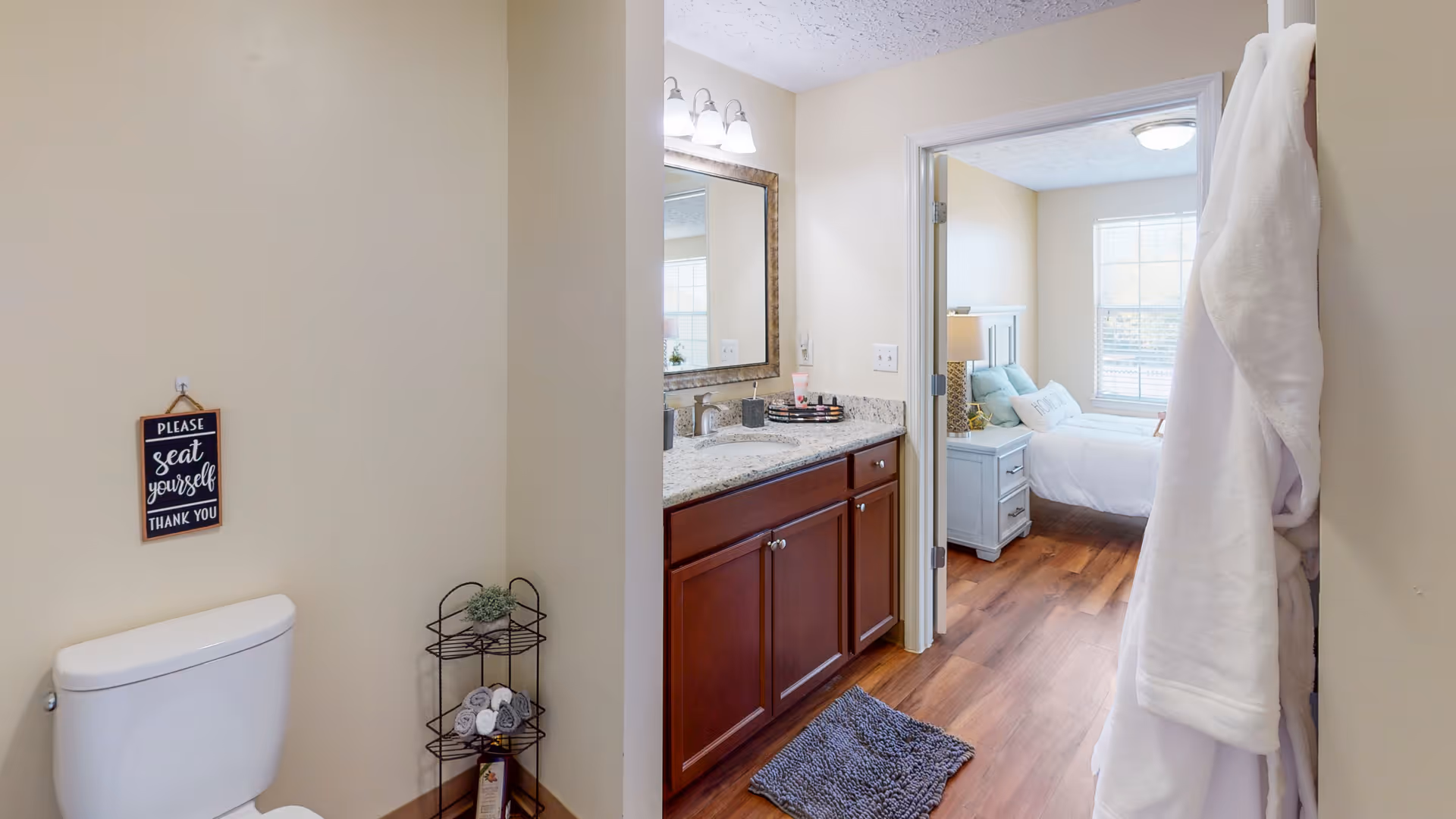 Bathroom with a toilet, granite-topped vanity and mirror, a robe hanging on the door, and a view into a bedroom.