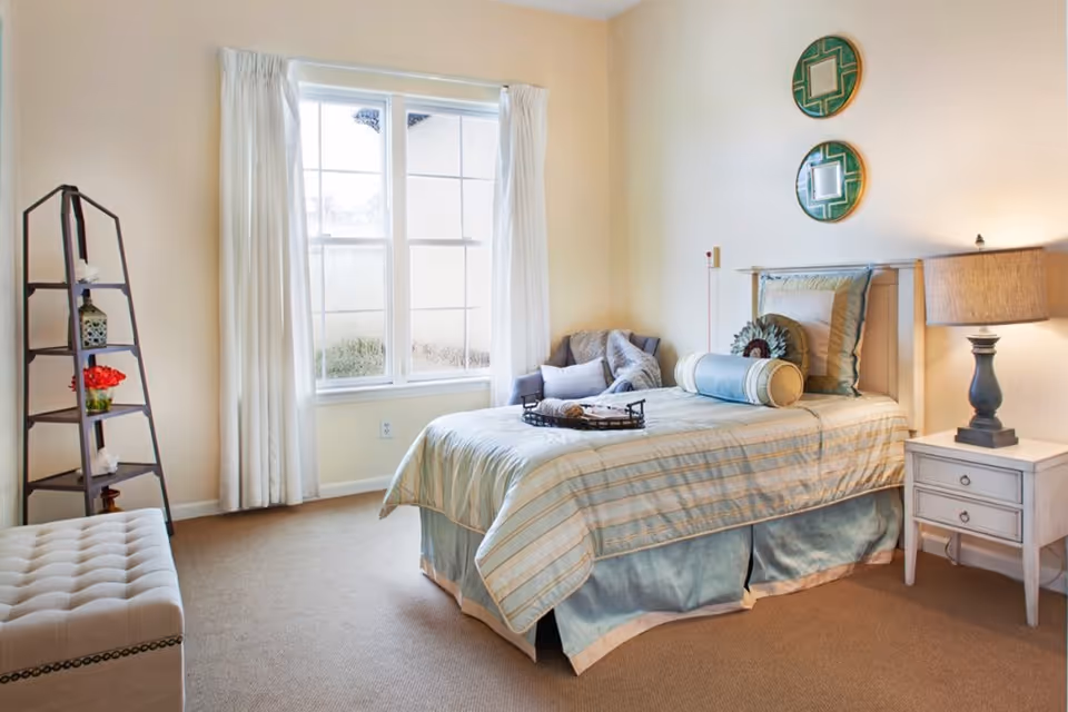 A cozy bedroom with a single bed dressed in light blue and beige striped bedding, a small white nightstand with a lamp, two decorative round mirrors on the wall, a window with white curtains, a gray armchair with pillows and a blanket, a tall dark wooden shelf with decorative items, and a beige cushioned bench.