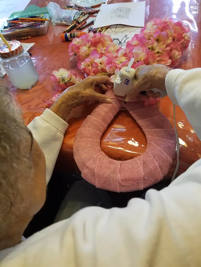An elderly person crafting a decorative wreath by attaching pink and white artificial flowers to a pink fabric-wrapped circular base using a hot glue gun. The table is covered with an orange tablecloth and scattered with crayons, glue sticks, and coloring sheets.