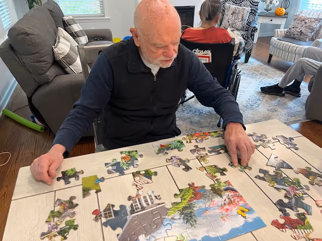 An elderly man is sitting at a table working on a colorful jigsaw puzzle in a cozy living room. Behind him, two other elderly individuals are seated, one in a wheelchair and the other on a chair. The room has comfortable seating including a sofa and armchairs, a patterned rug, and a small table with a pumpkin decoration.