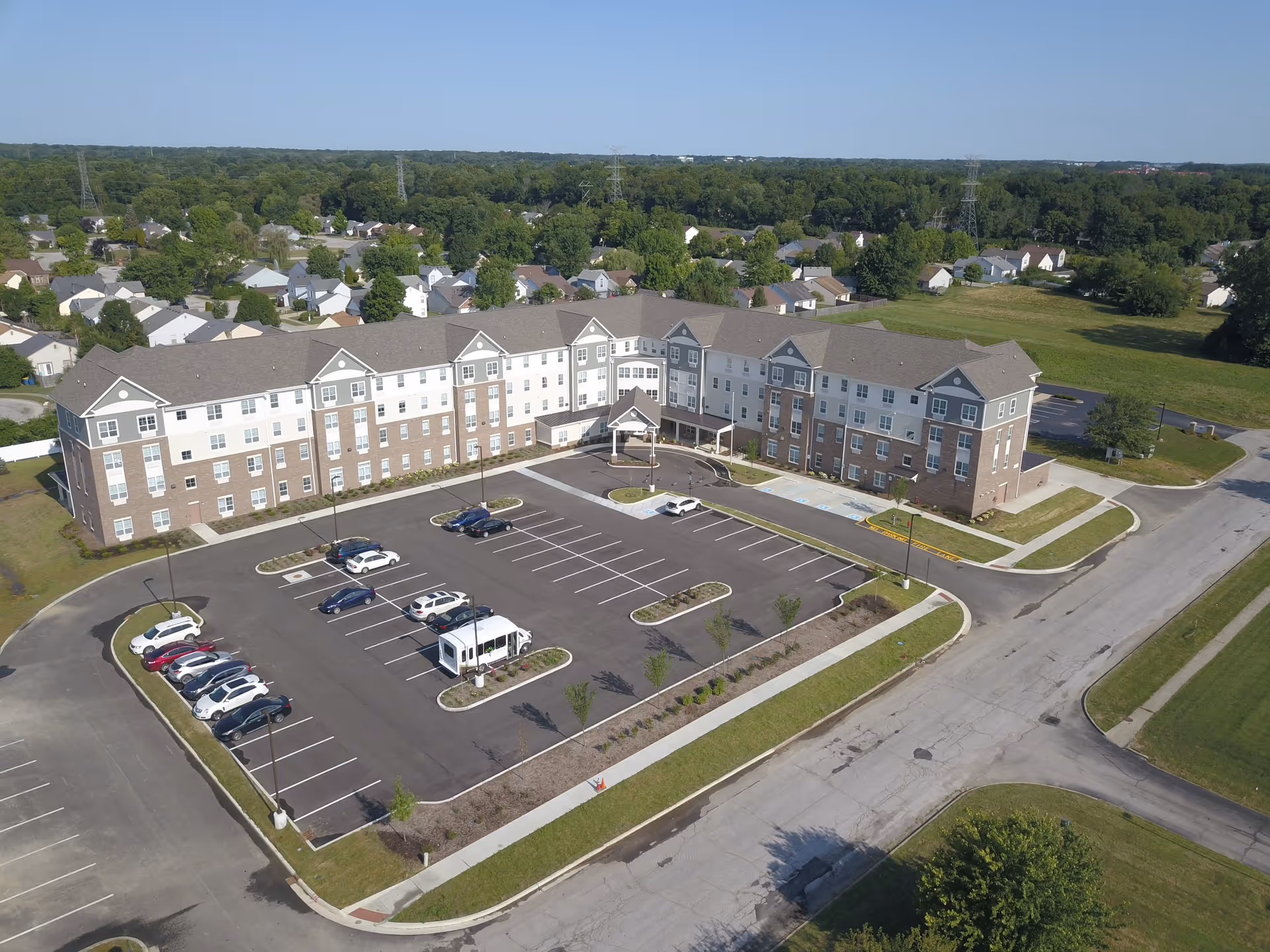 Aerial view of a large, three-story senior living facility named Oasis at 56th, surrounded by a parking lot with several cars parked. The building has a combination of brick and light-colored siding exterior, with multiple windows and a covered entrance. The facility is located in a suburban area with houses and greenery in the background under a clear blue sky.