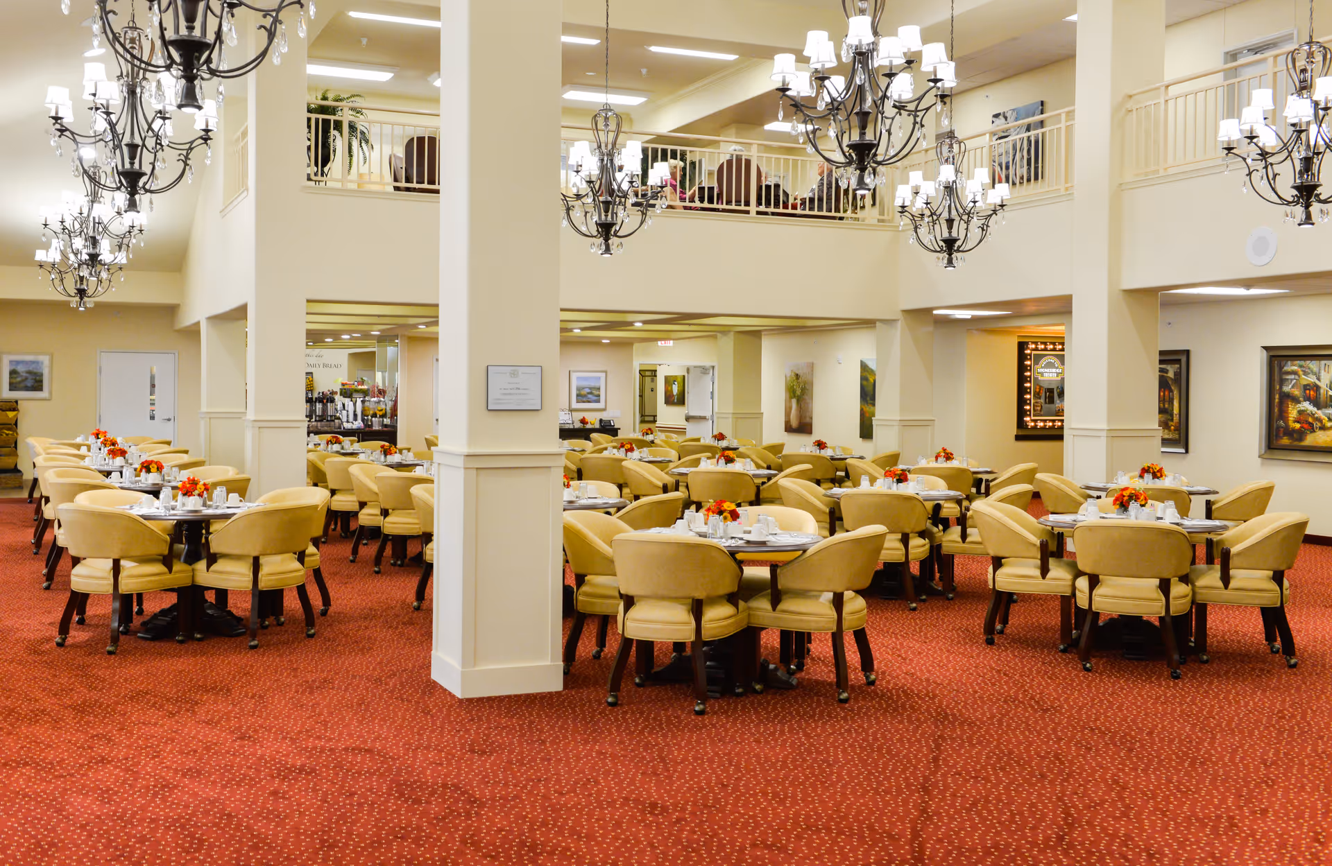 Spacious dining room with multiple round tables surrounded by beige upholstered chairs, each table set with white dishes and small floral centerpieces. The room features red carpet flooring, cream-colored walls, large chandeliers hanging from the ceiling, and a second-floor balcony overlooking the area.