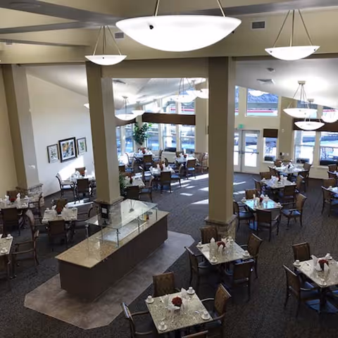Spacious senior living dining area with multiple set tables, a central serving counter, and large pendant lights.