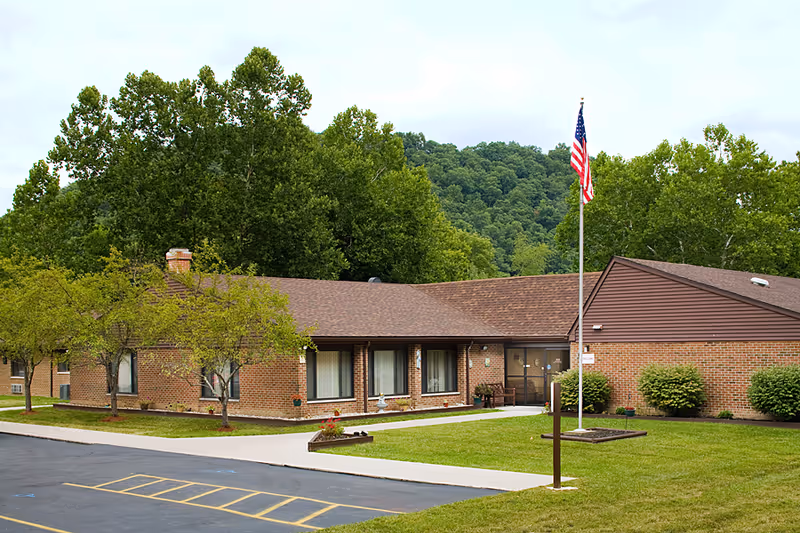 Single-story brick building with a brown roof, surrounded by green trees and grass. There is a paved parking area with yellow markings in front, a flagpole with an American flag, and a sidewalk leading to the entrance of the building.