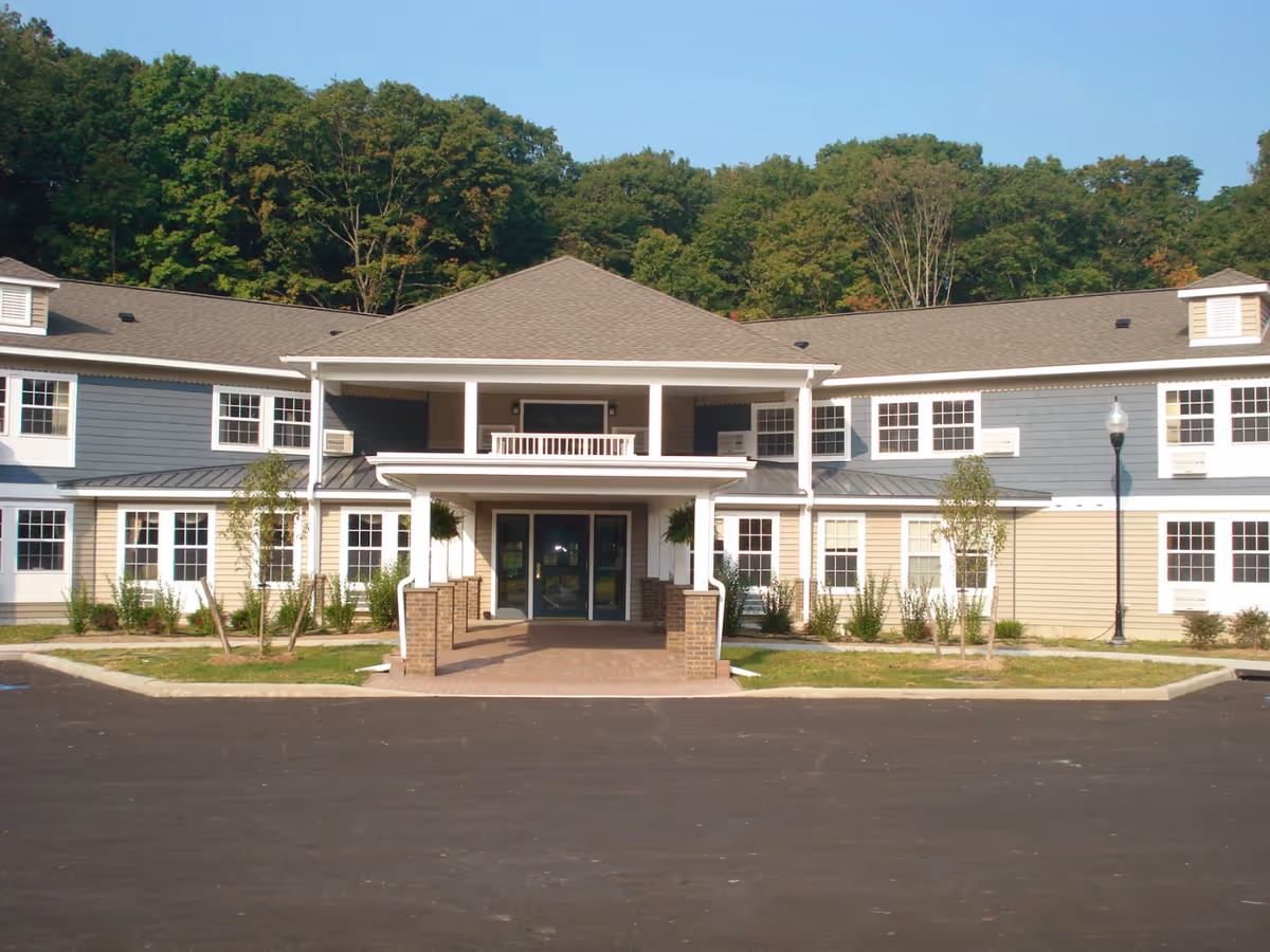 Front exterior view of a two-story senior living facility building named Mountain Valley Manor Adult Home, featuring a covered entrance with brick pillars, multiple windows, and surrounded by greenery and trees in the background.