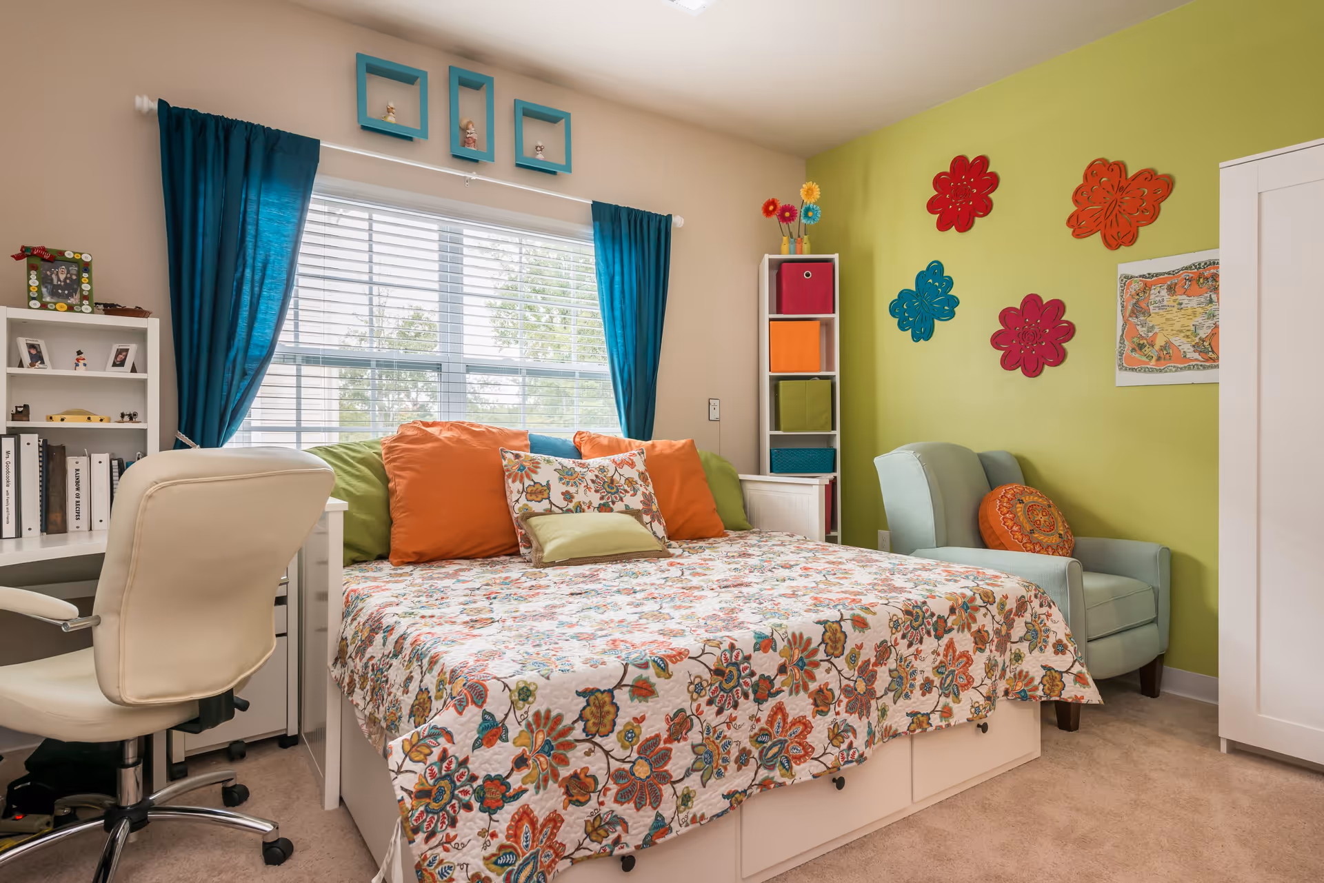 A bright and colorful bedroom featuring a bed with a floral patterned bedspread and multiple pillows in orange, green, and floral designs. To the left of the bed is a white desk with a white office chair and a small white bookshelf with books and framed photos. The window behind the bed has white blinds and teal curtains. On the right side of the room, there is a light green armchair with an orange decorative pillow, a white storage unit with colorful fabric bins, and a white wardrobe. The walls are painted beige and light green, decorated with colorful flower-shaped wall hangings and a framed map.