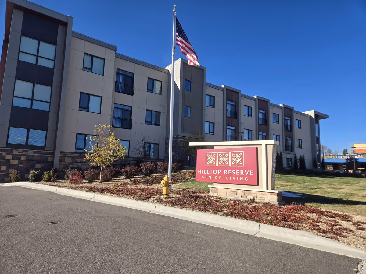 Front exterior of the Hilltop Reserve Senior Living building with its sign, flagpole, and landscaped grounds under a clear blue sky.