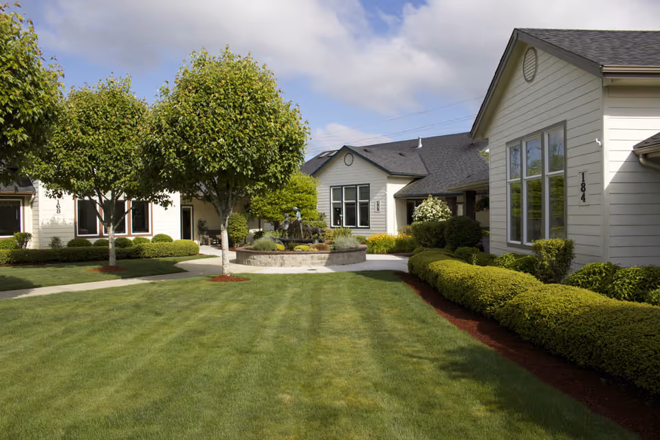 A well-maintained outdoor garden area at Gateway Gardens featuring neatly trimmed green grass, manicured bushes, and several small trees. There are light-colored buildings with large windows surrounding the garden, and a circular stone planter with a small fountain or statue in the center. The sky is partly cloudy with blue patches visible.