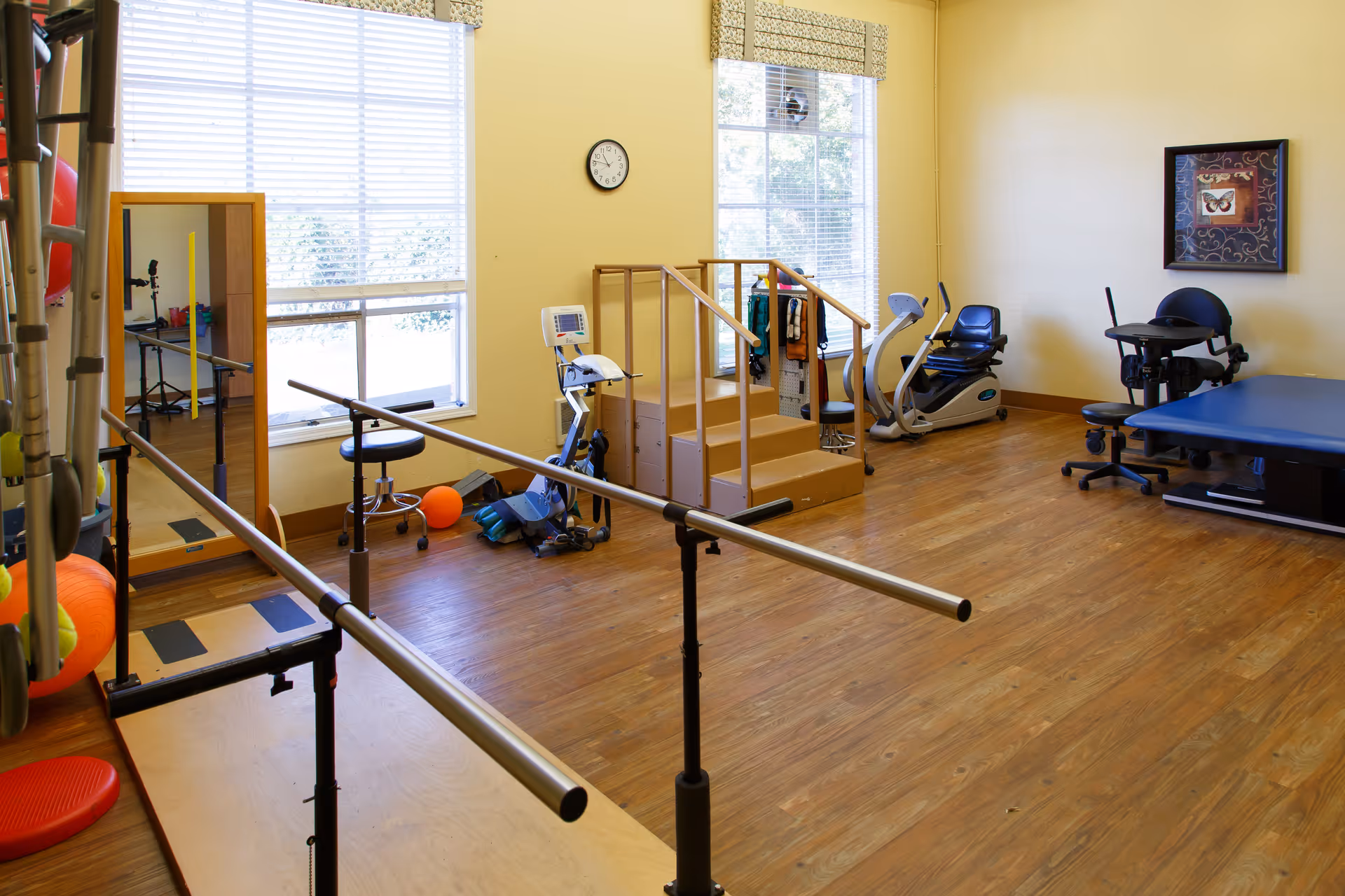 A physical therapy room with wooden floors and yellow walls, featuring parallel bars for walking exercises, a small set of stairs with handrails, an exercise bike, a therapy table, a chair with wheels, a large mirror, and various therapy equipment and exercise balls. Two large windows with blinds allow natural light into the room.
