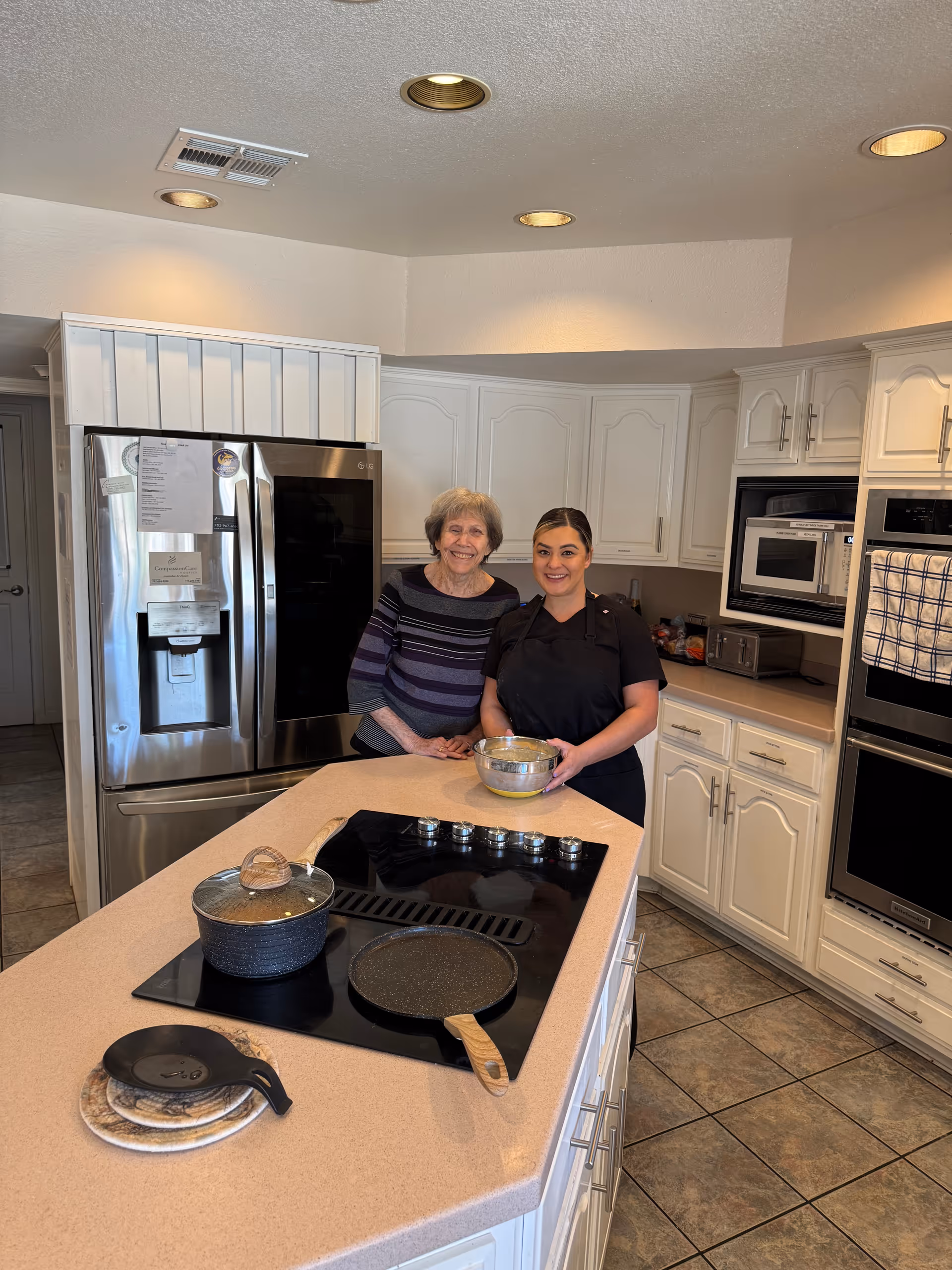 An elderly woman and a younger woman standing together in a bright kitchen. The elderly woman is wearing a striped sweater and the younger woman is dressed in a black chef's outfit holding a mixing bowl. The kitchen features white cabinets, a stainless steel refrigerator, a stovetop with a pot and pan, and a tiled floor.