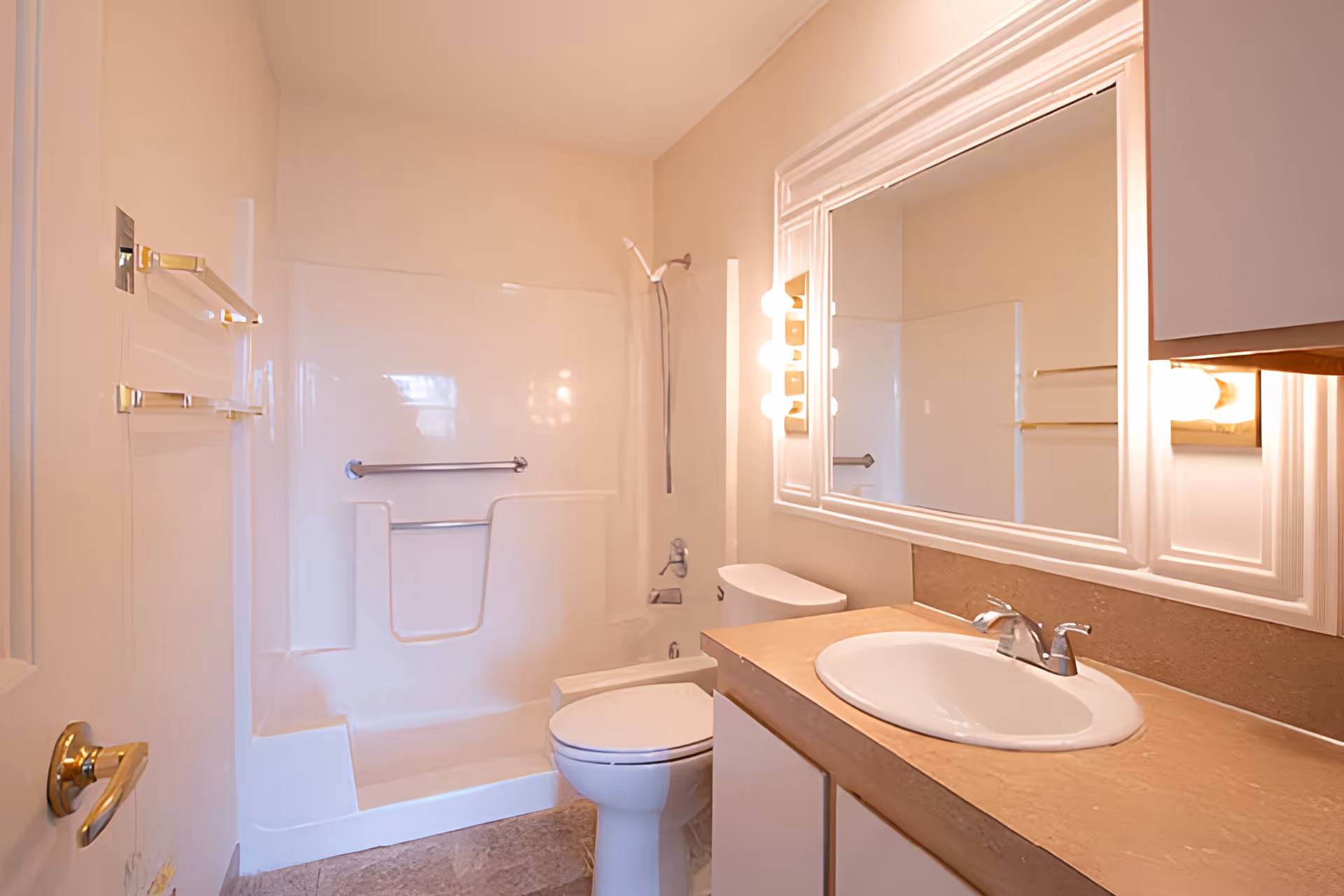 A clean bathroom featuring a white bathtub with a showerhead and grab bars, a white toilet, a sink with a beige countertop, a large mirror with white framing, and wall-mounted lights beside the mirror.