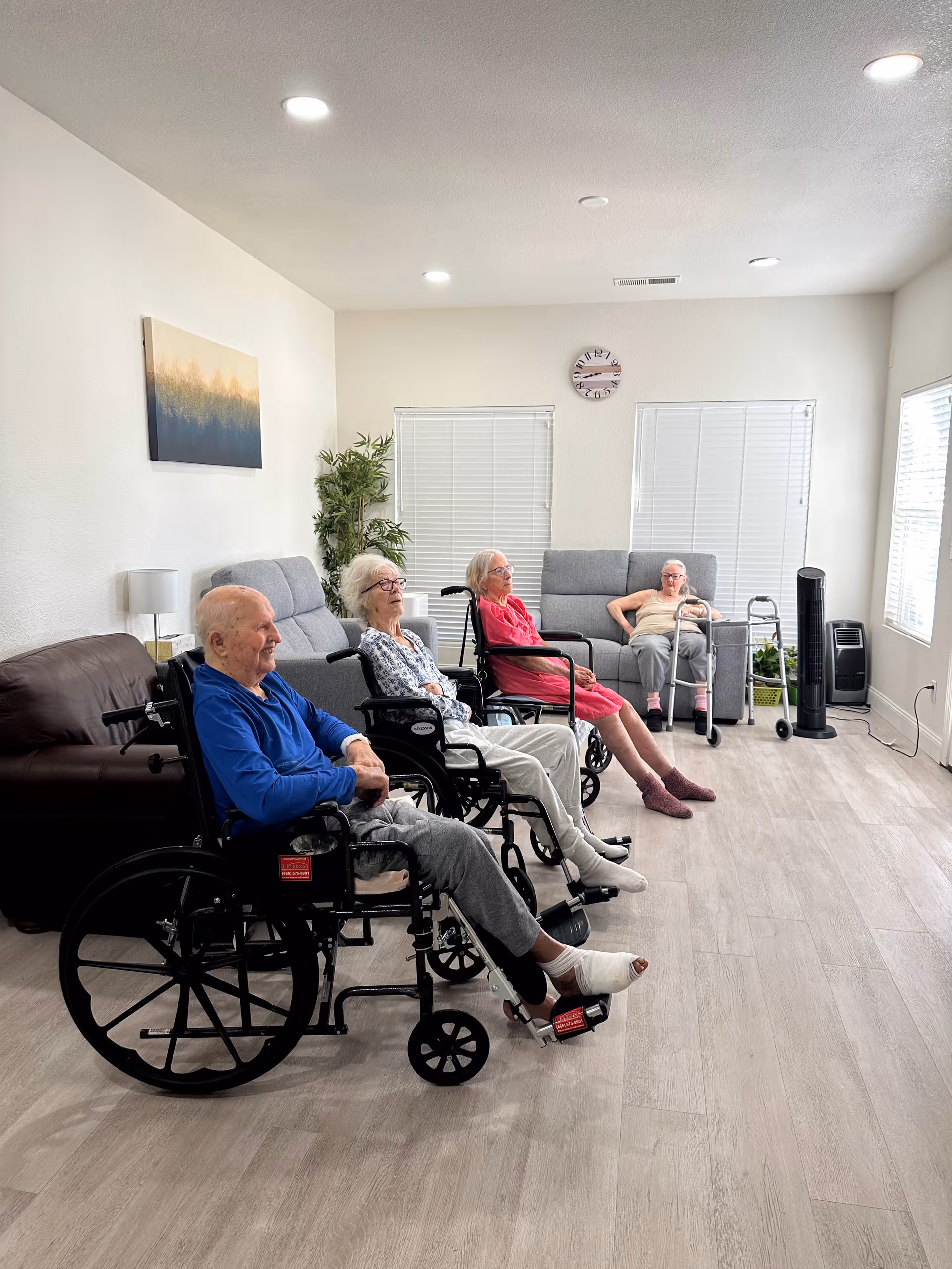 A bright living room with four elderly individuals seated, three in wheelchairs and one on a sofa with a walker nearby. The room has light-colored walls, wood flooring, a painting on the wall, two windows with blinds, a clock, a potted plant, and a standing fan.
