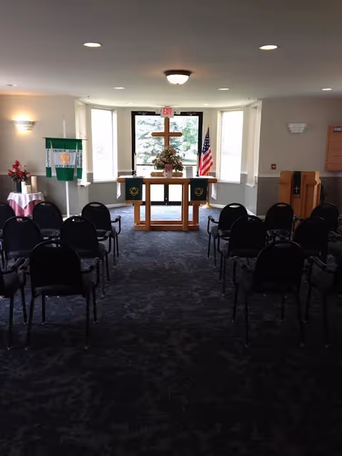 Interior view of a small chapel or worship room with rows of black chairs facing a wooden altar adorned with a cross, flowers, and religious banners. An American flag is positioned to the right of the altar, and windows behind the altar allow natural light to enter the room.