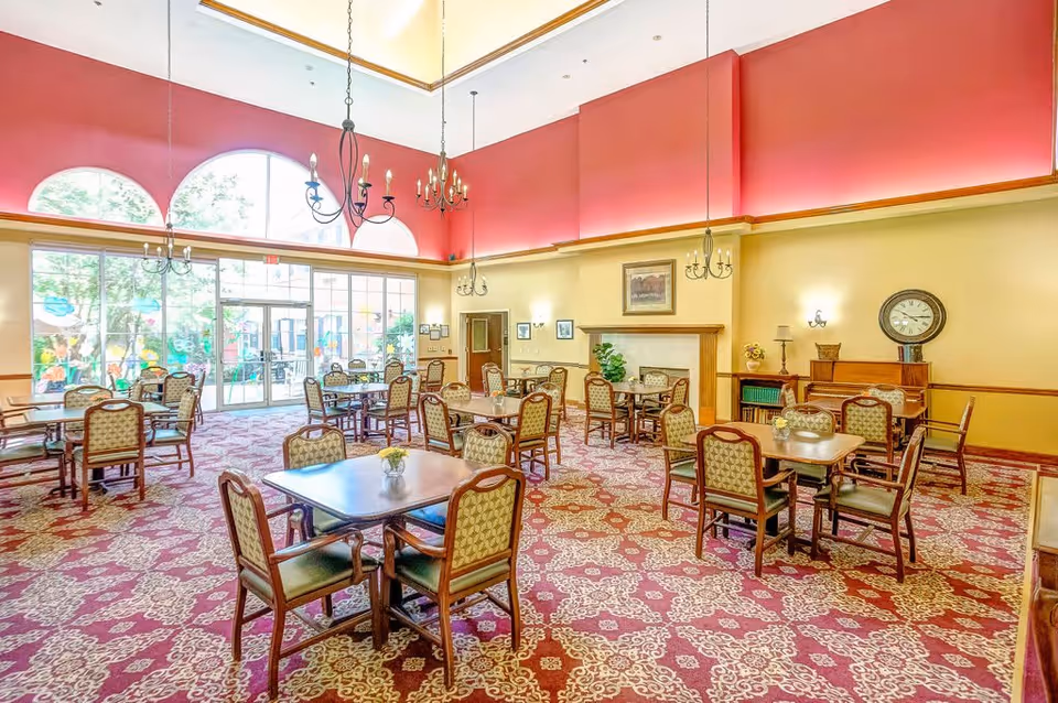 Spacious senior living dining room with multiple tables and chairs, chandeliers, patterned carpet and large arched windows to an outdoor area.