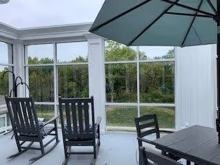 A bright enclosed patio area with large windows overlooking greenery. The space features two black rocking chairs, a black table with matching chairs, and a large green umbrella providing shade.